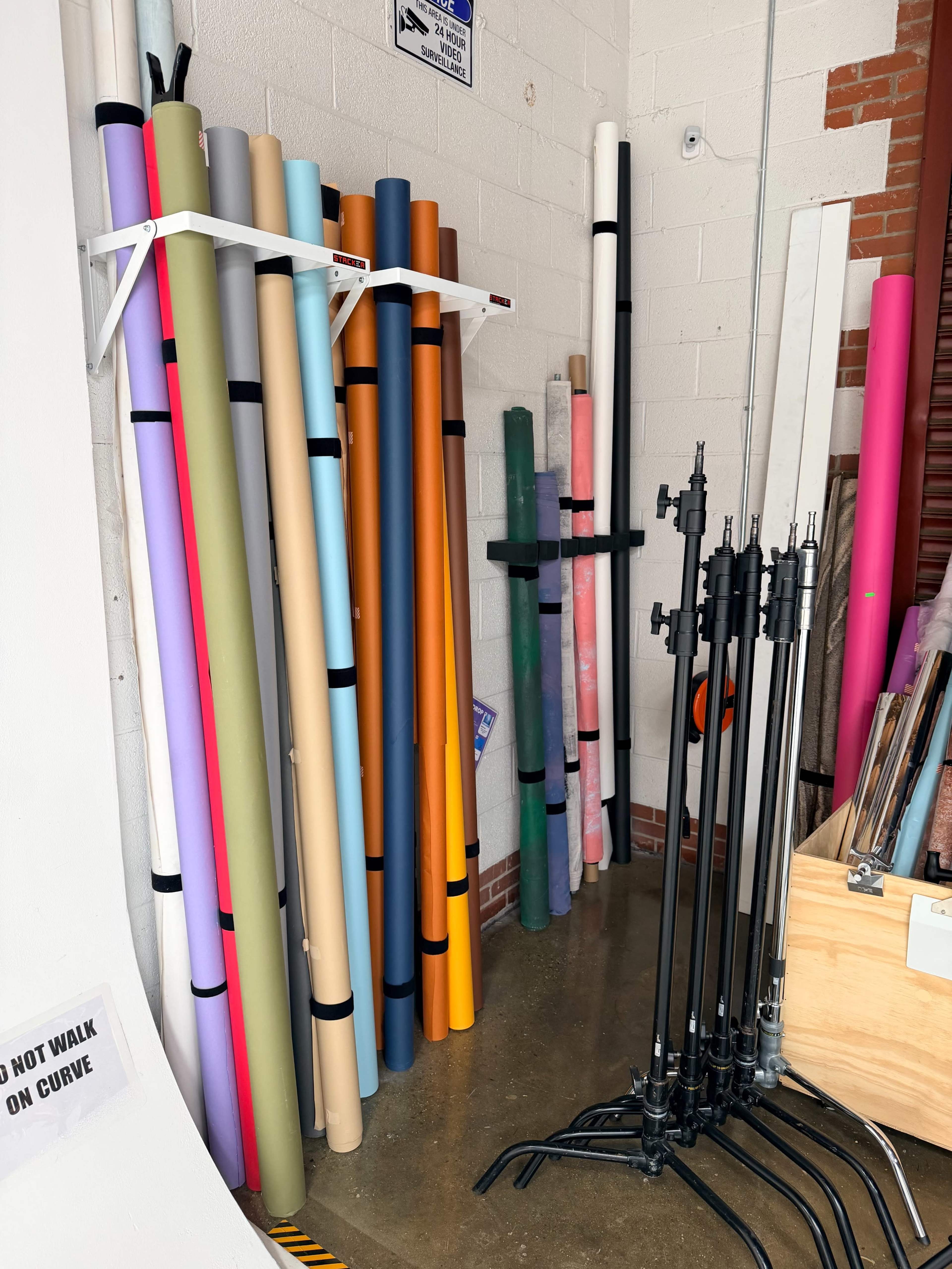 The image shows a storage area with rolls of colorful paper and several black stands against a wall.