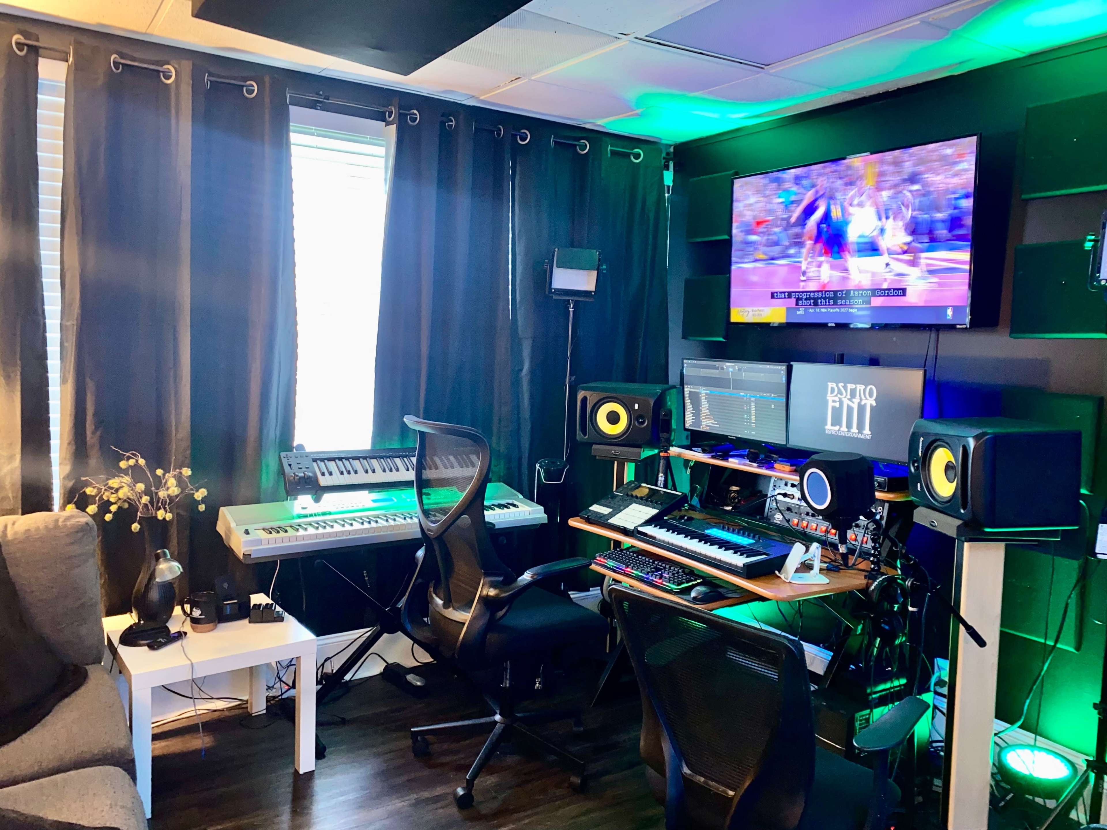 The image shows a music production studio featuring a keyboard, mixing equipment, and a television, with dark curtains and colorful lights enhancing the ambiance.