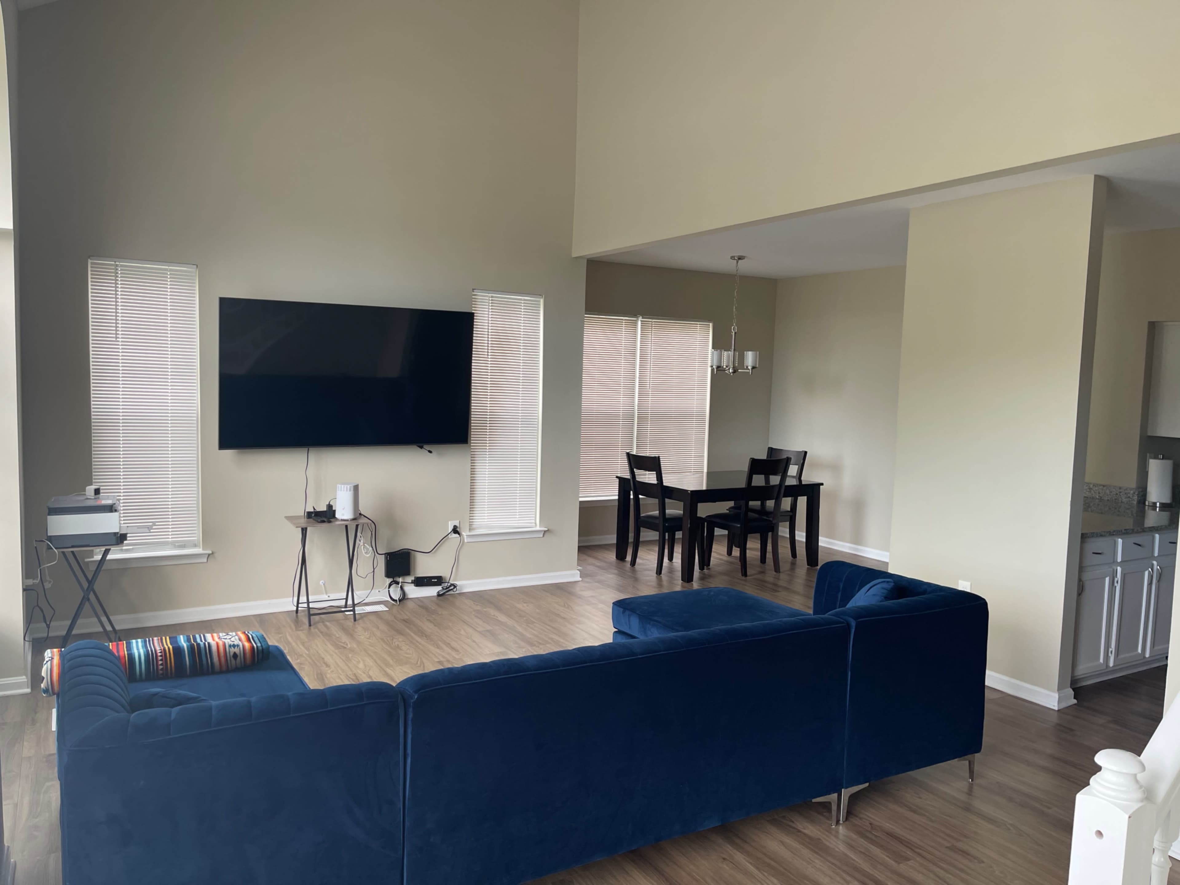 The image shows a spacious living area with a blue sectional sofa, a mounted television on the wall, a dining table with black chairs, and large windows with blinds.