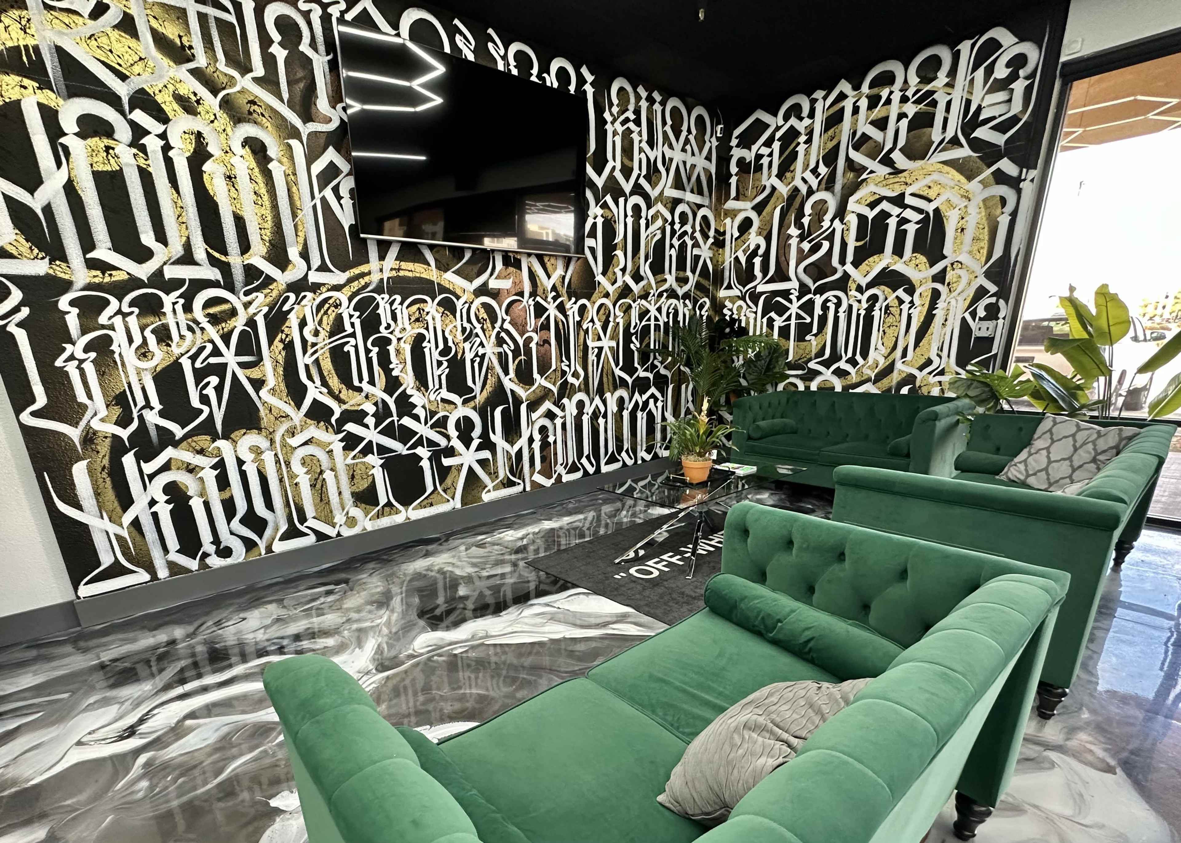 A modern lounge area with green velvet couches facing a wall covered in bold, artistic black and gold typography and a mounted television above.