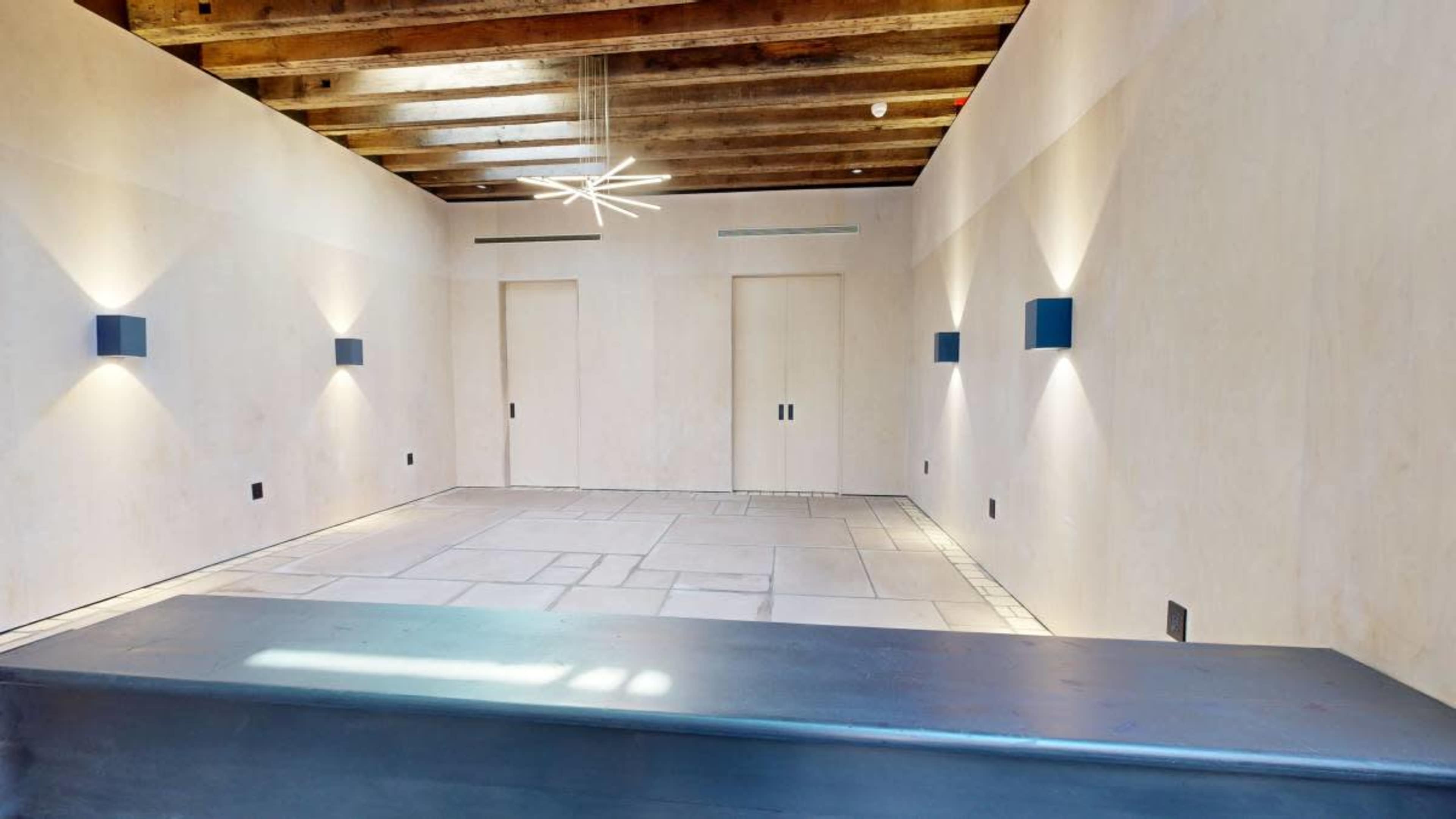 The image shows a minimalistic interior room with exposed wooden beams, two light fixtures on the walls, and a stone floor.