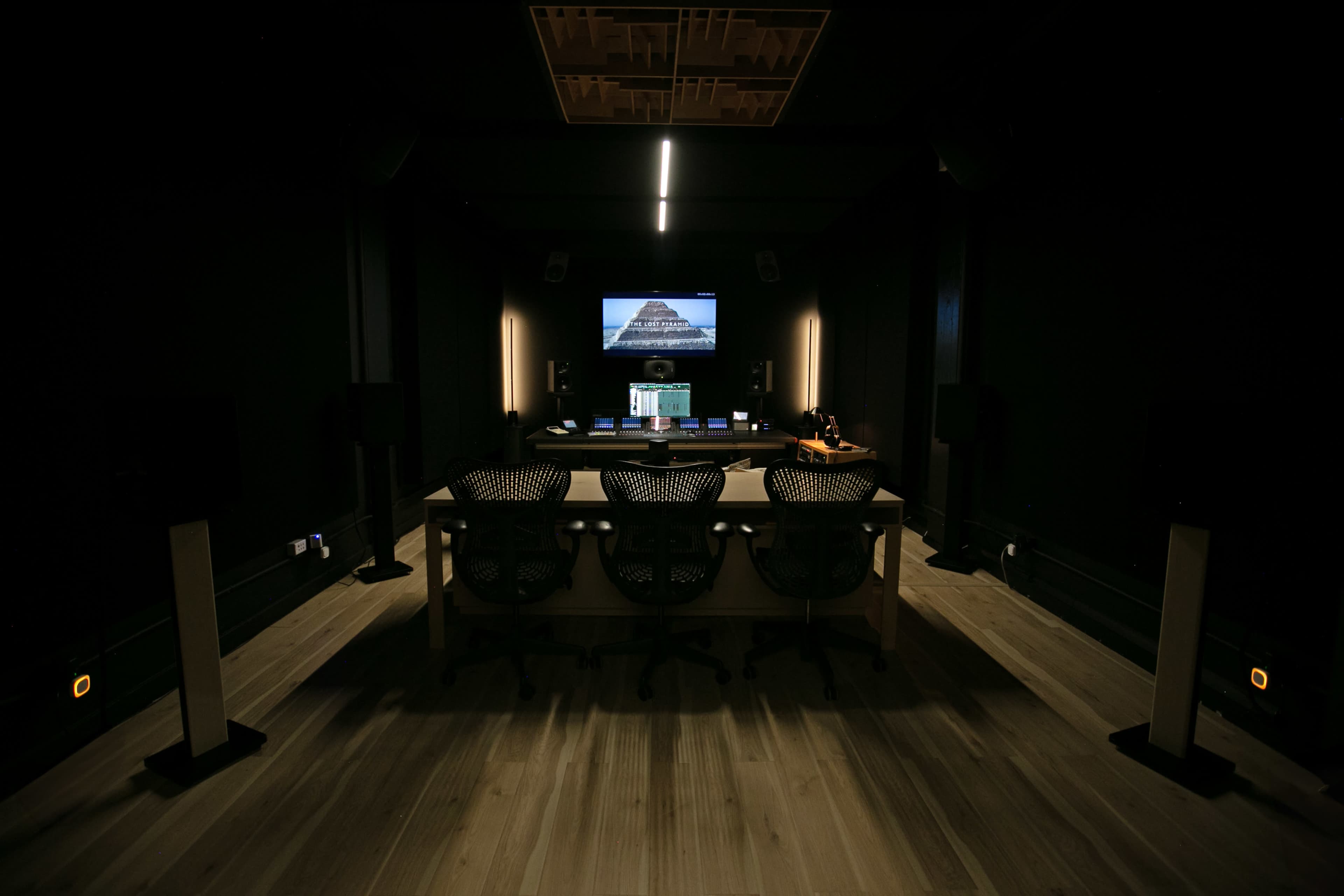 A dimly lit recording studio with three chairs facing a large screen and audio equipment.