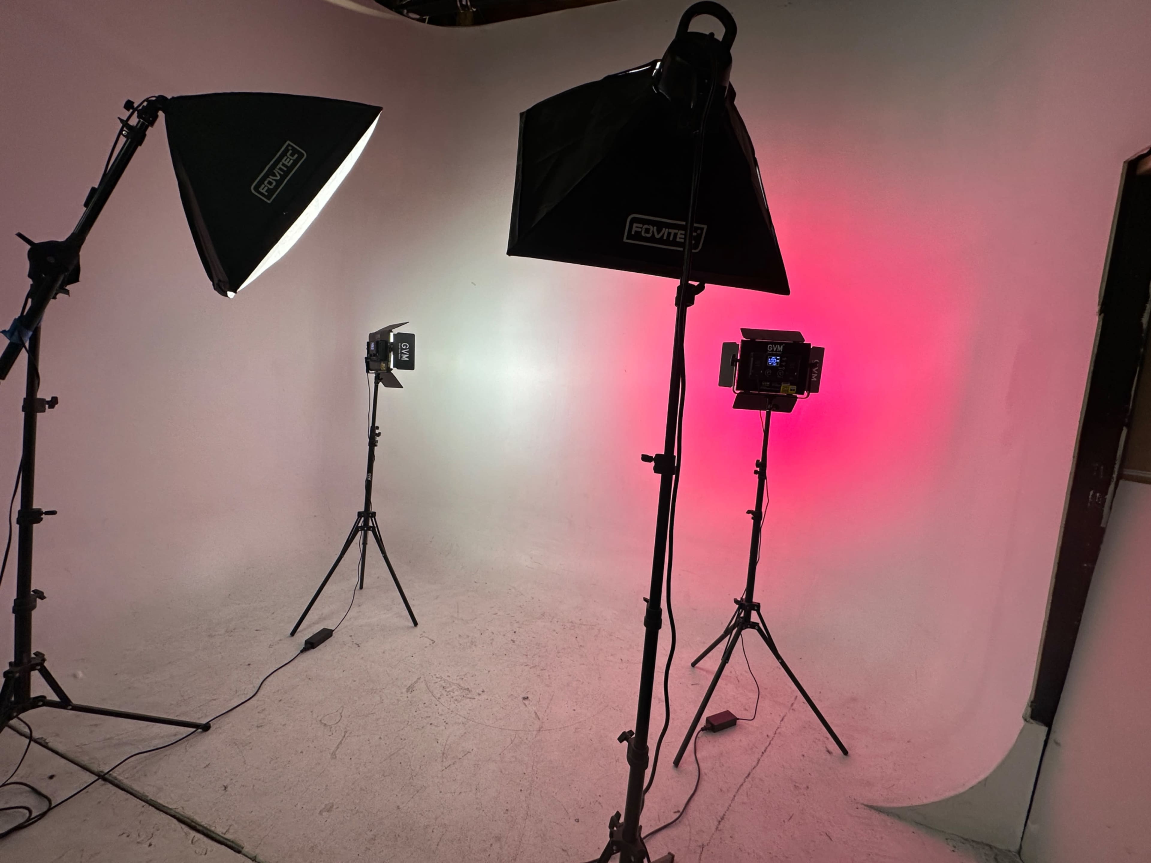 The image shows a photography studio setup with multiple lighting equipment positioned around a smooth, light-colored backdrop.