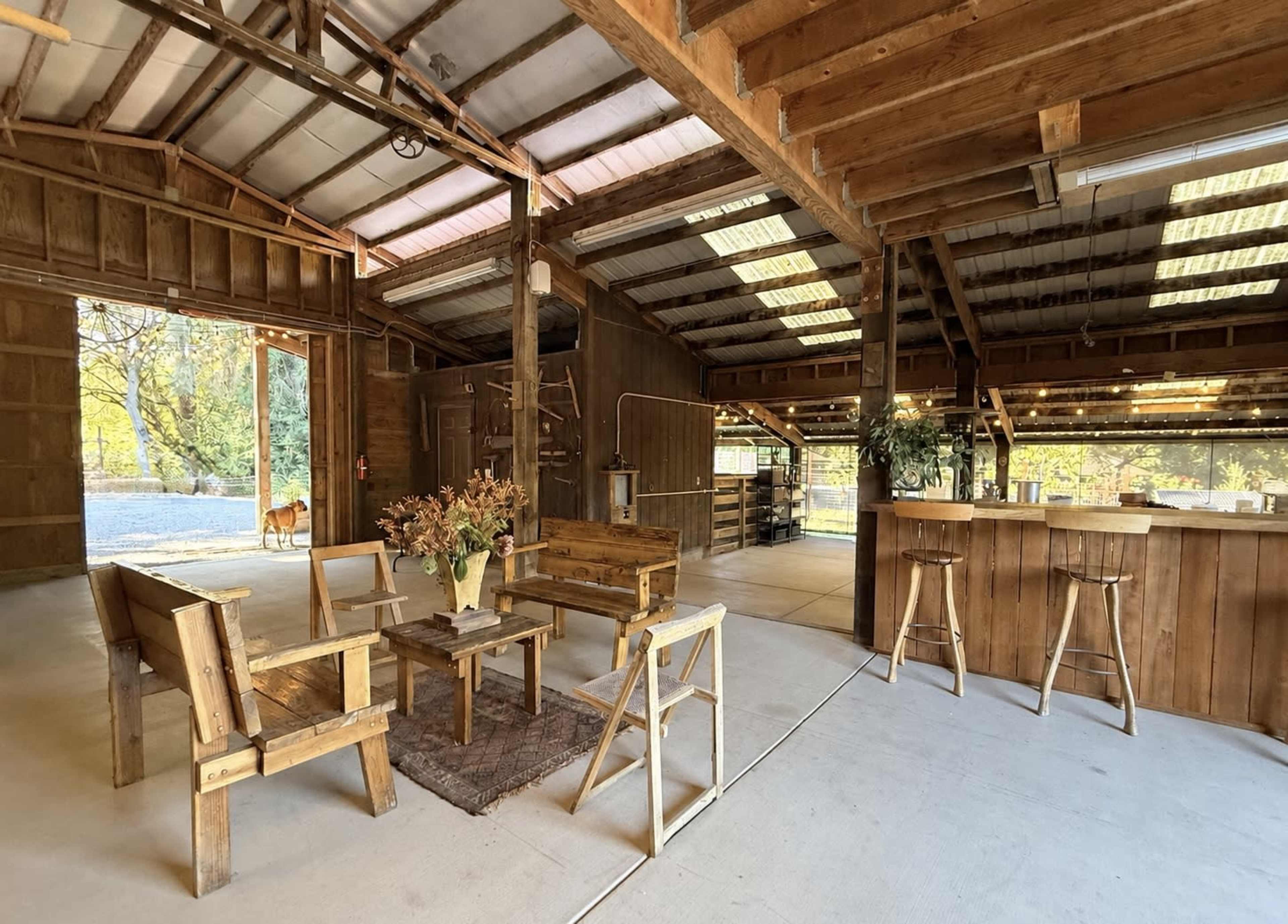 The image shows a spacious wooden barn interior with seating arrangements, a bar area, and an open entrance revealing a dog outside.
