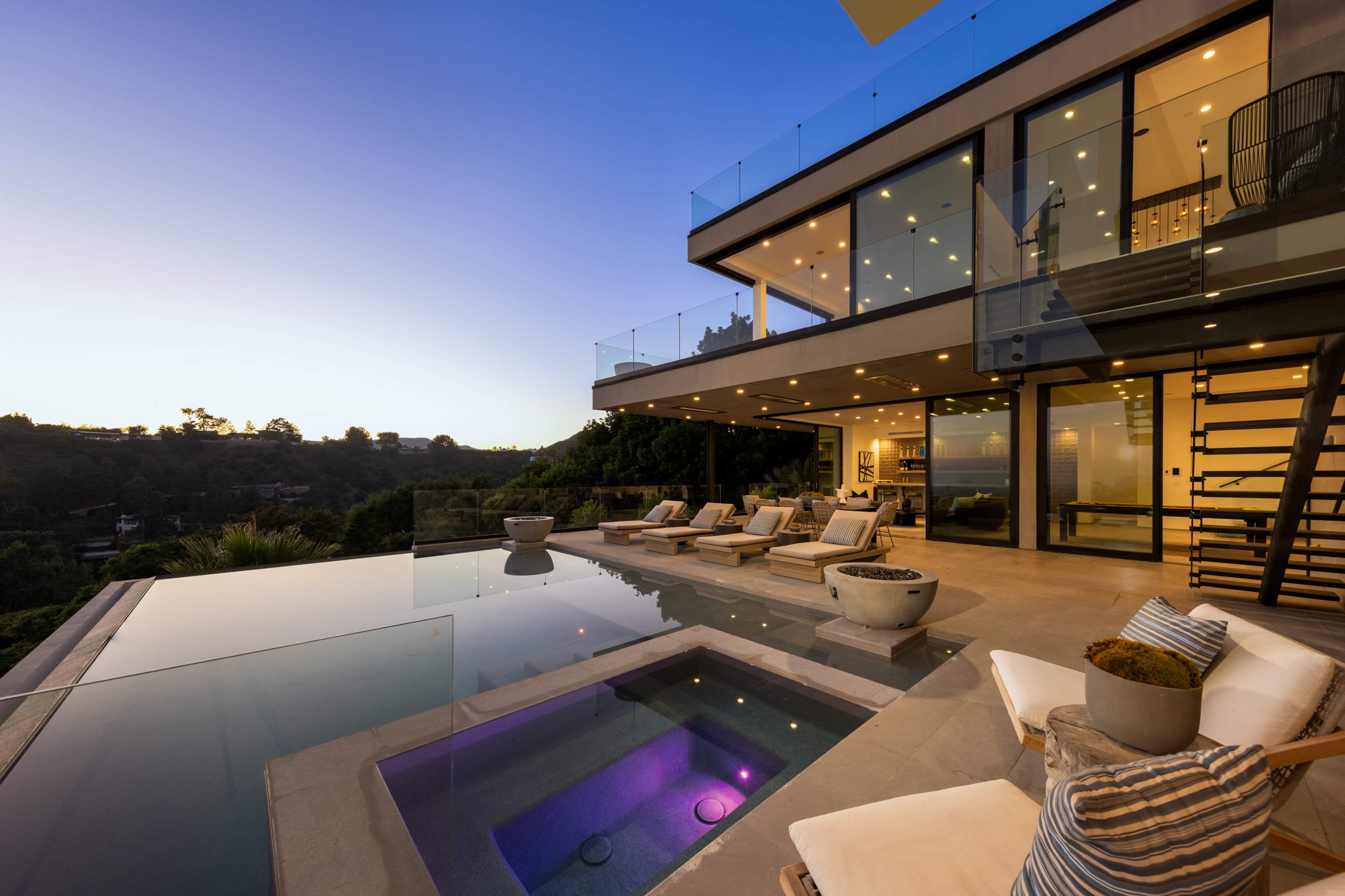 A modern house with large glass walls overlooks a swimming pool and spa at dusk.
