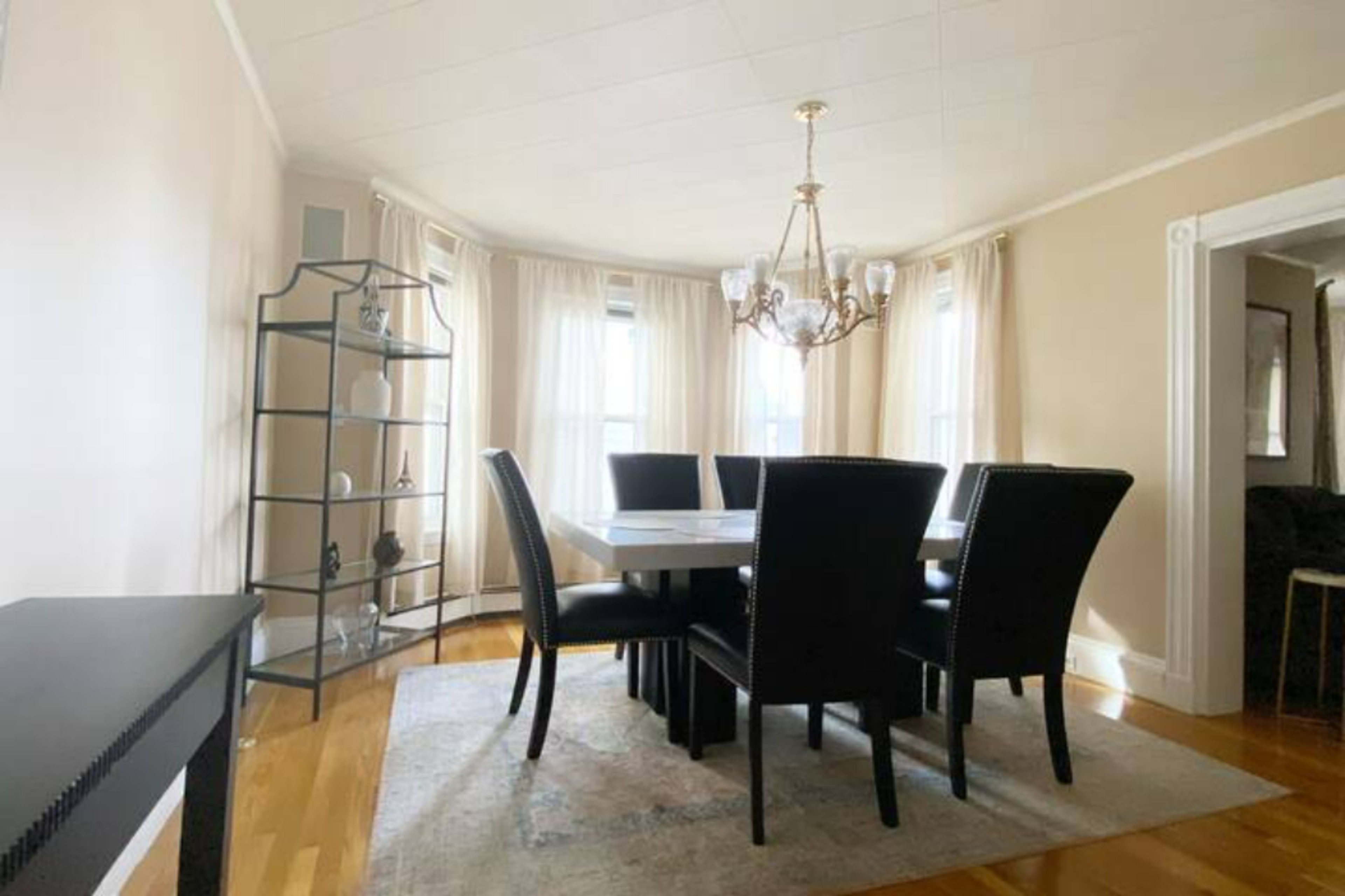 A dining room features a rectangular table surrounded by six black chairs, with a chandelier above and a tall shelving unit alongside the windows.