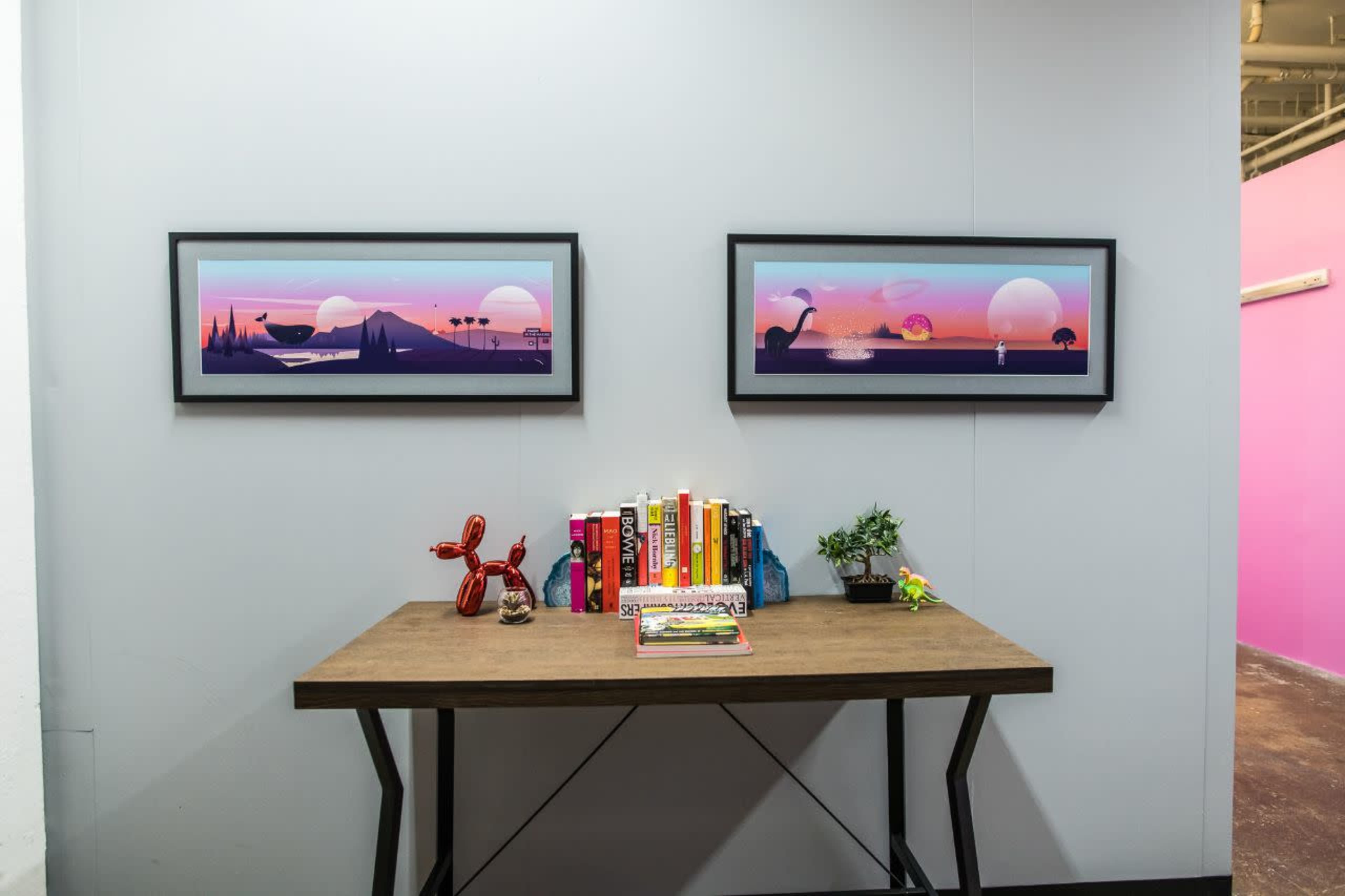A wooden table displaying books, a small plant, and a balloon animal sculpture, with two framed panoramic artworks hung on the wall above it.