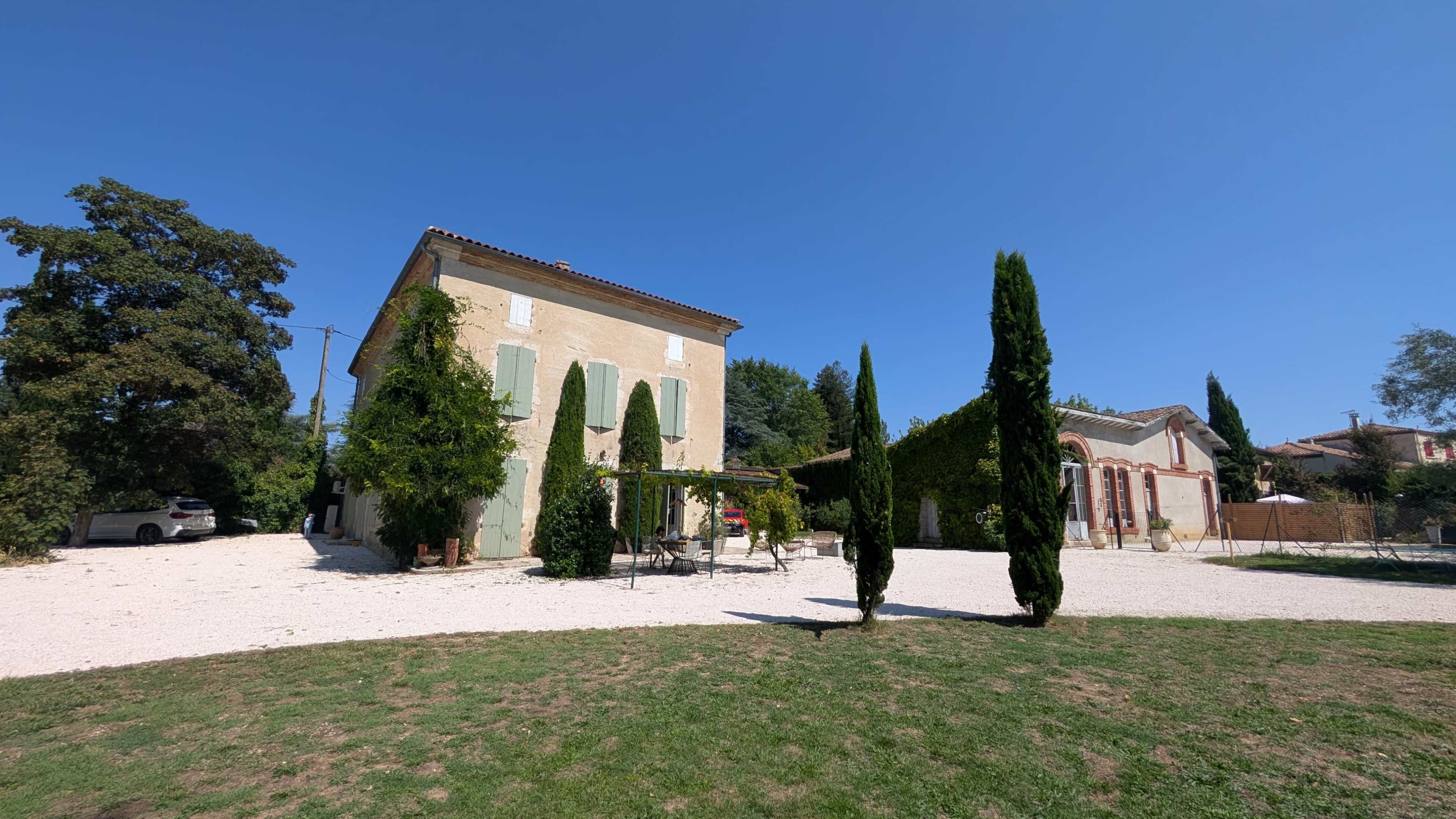The image shows a two-story light-colored house with green shutters surrounded by neatly trimmed cypress trees and a gravel area under a clear blue sky.