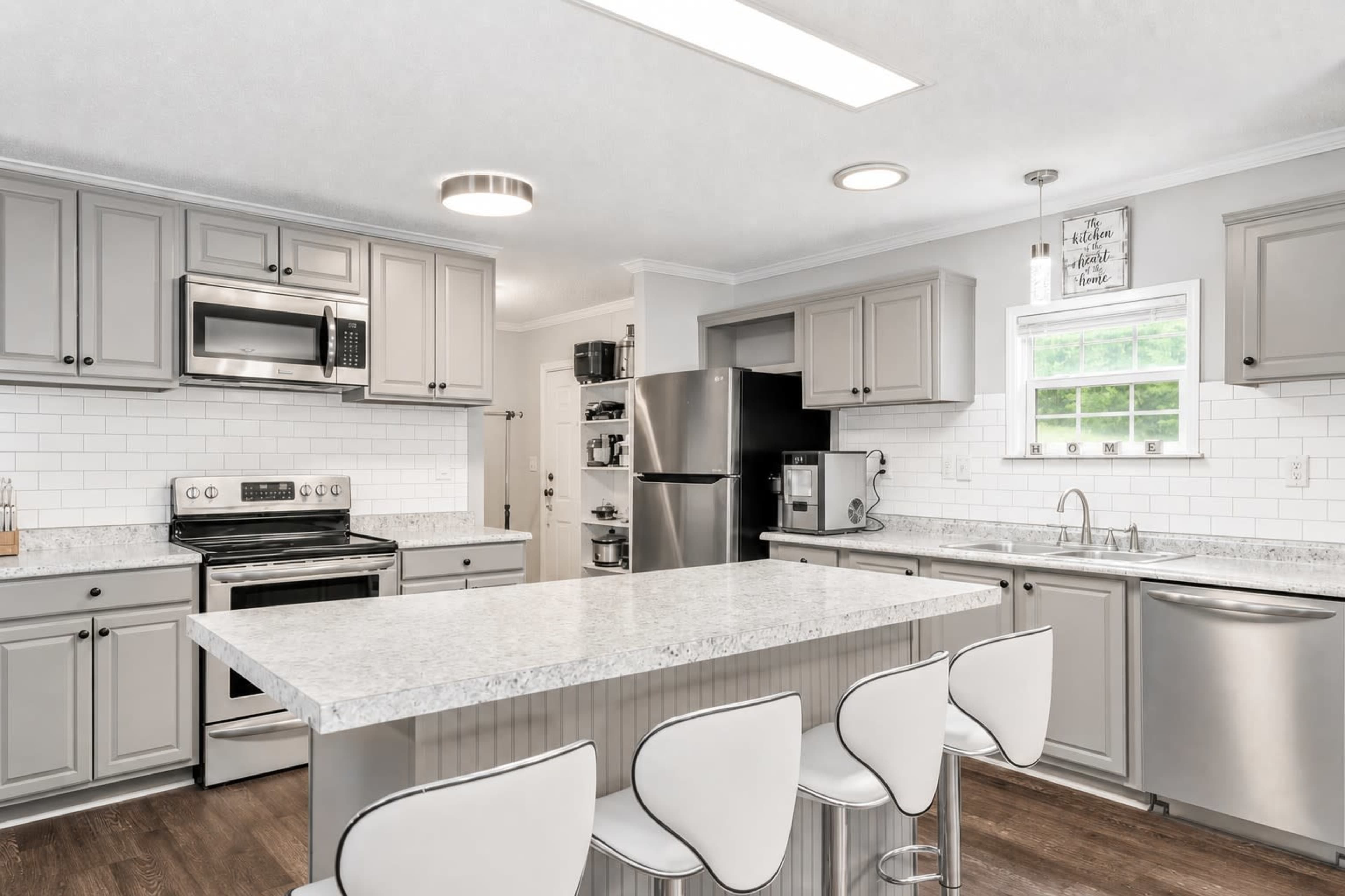 The image shows a modern kitchen featuring gray cabinetry, a spacious island with white bar stools, stainless steel appliances, and a marble countertop.