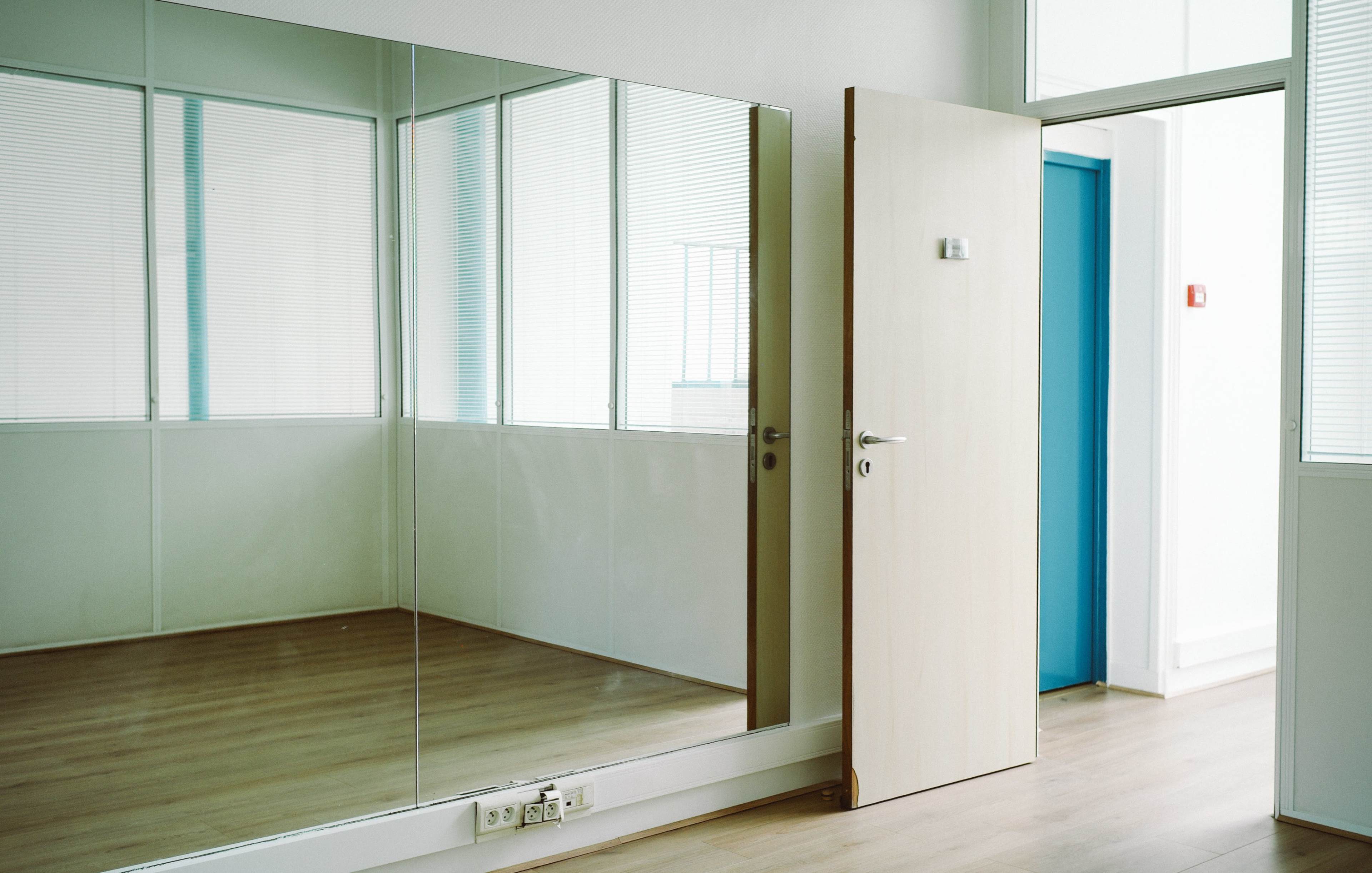 The image features a minimalist room with a large mirror wall, a wooden door partially open, and light-colored flooring.