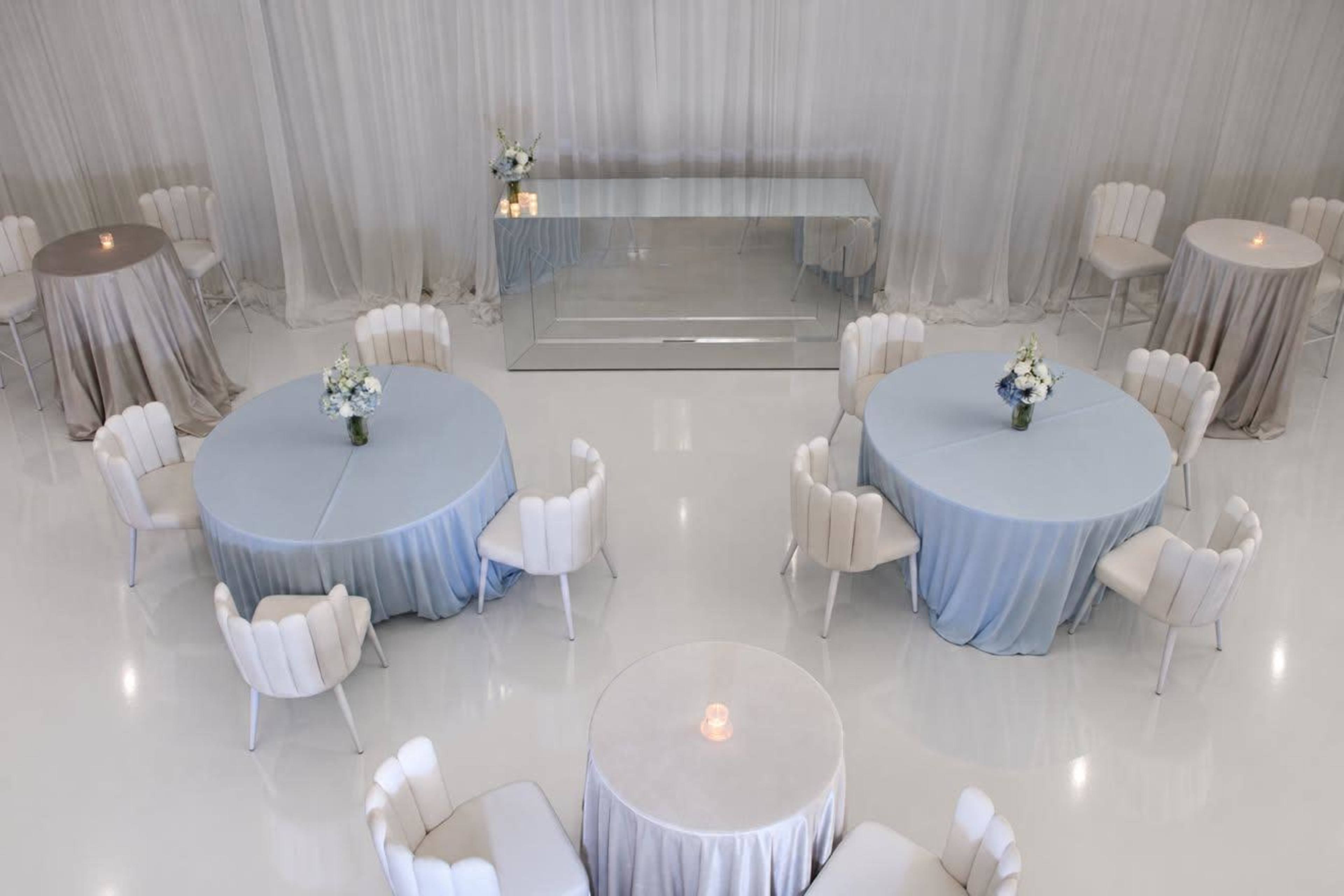 The image shows a modern event space with round tables covered in light blue and beige linens, surrounded by white chairs, and a mirrored centerpiece table at the front.