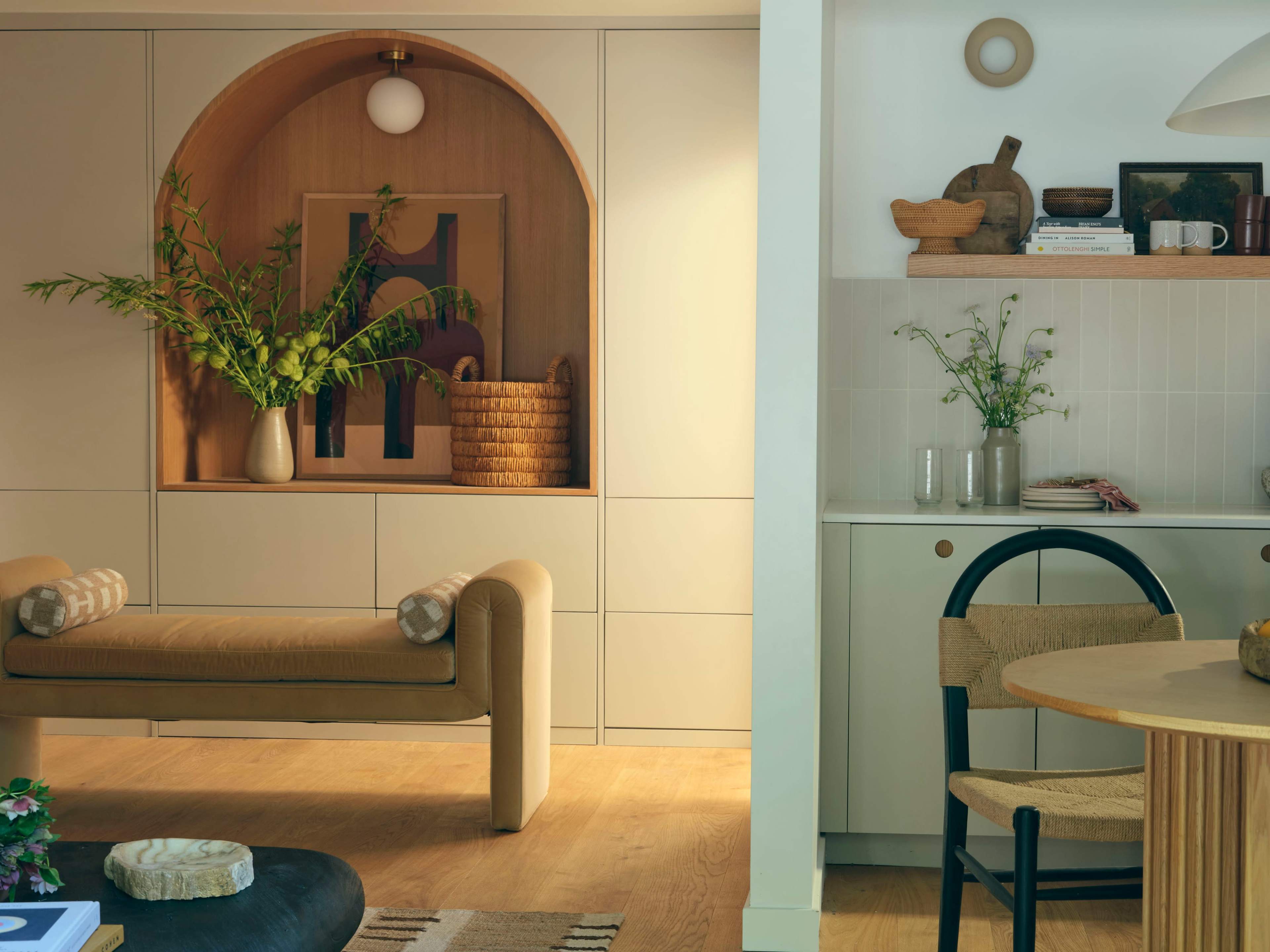 The image shows a modern interior space featuring a curved alcove with a decorative arrangement, a light-colored bench, and a dining area with a round table and woven chair.