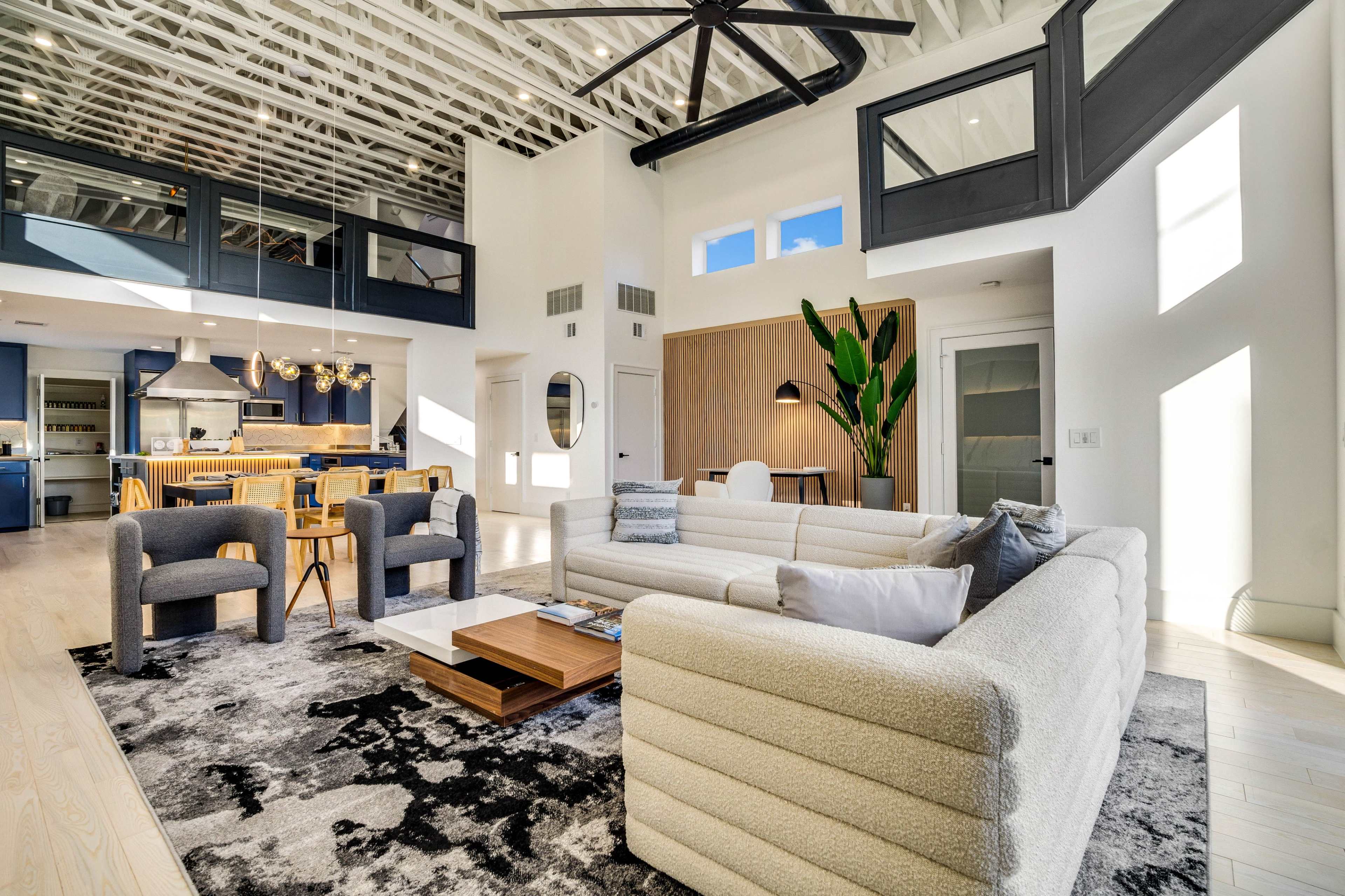 The living room features a spacious layout with a large sectional sofa, two accent chairs, a coffee table, and a tall indoor plant, all set on a patterned rug beneath a high ceiling with exposed beams.