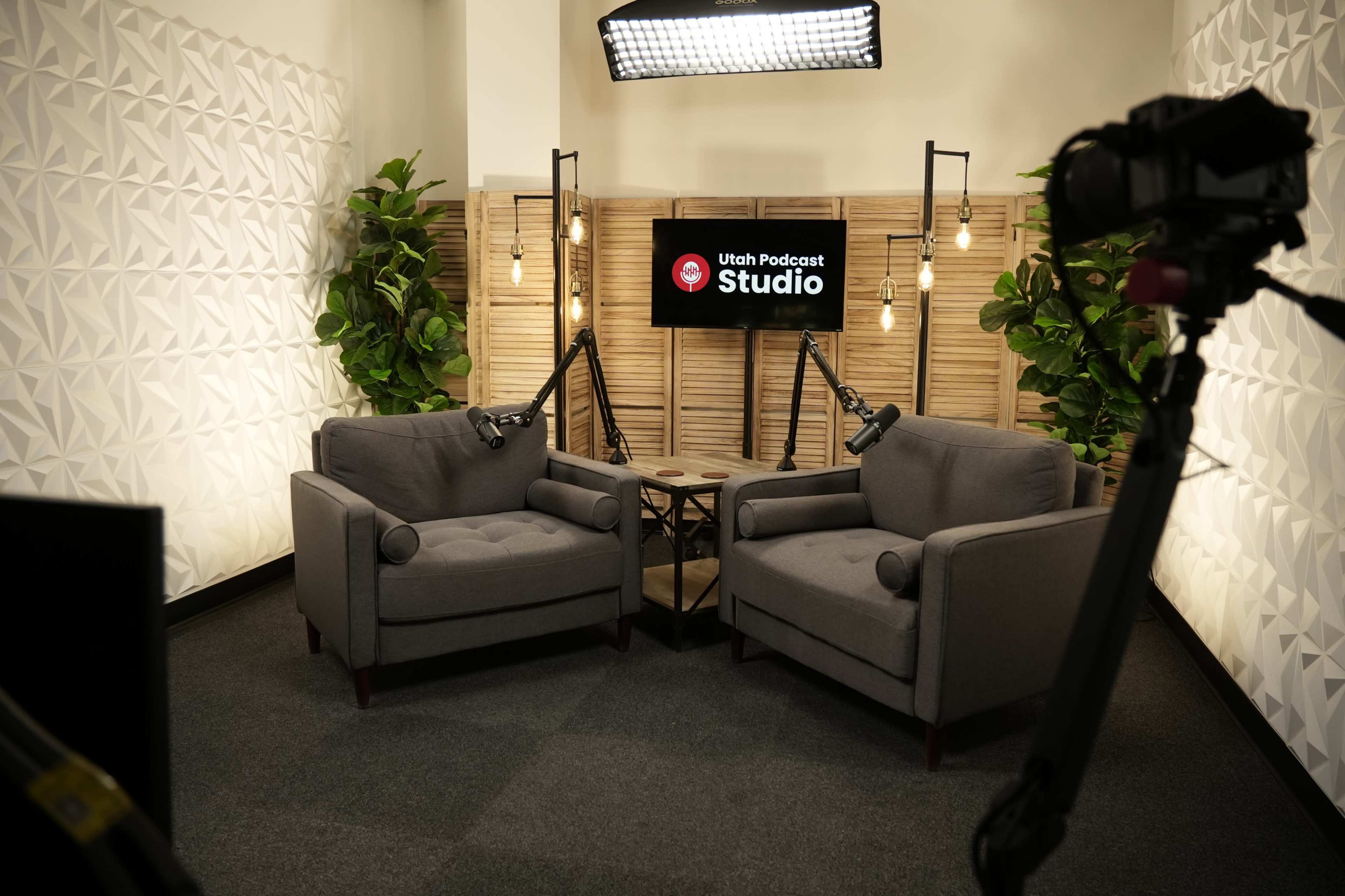 The image shows a recording studio setup featuring two gray armchairs facing each other, surrounded by decorative plants and lighting, with a screen displaying "Utah Podcast Studio" in the background.