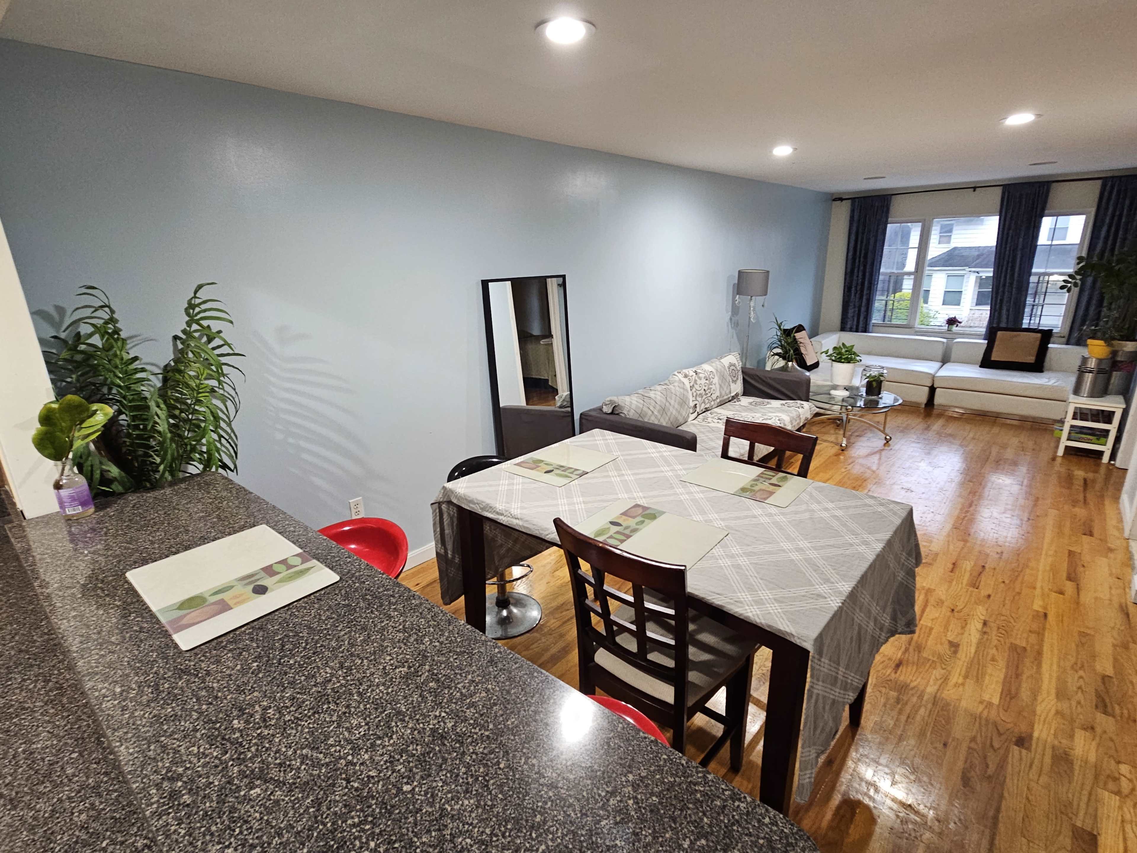 A spacious living area features a dining table with place settings, a mirror, a couch, and large windows in a well-lit setting.