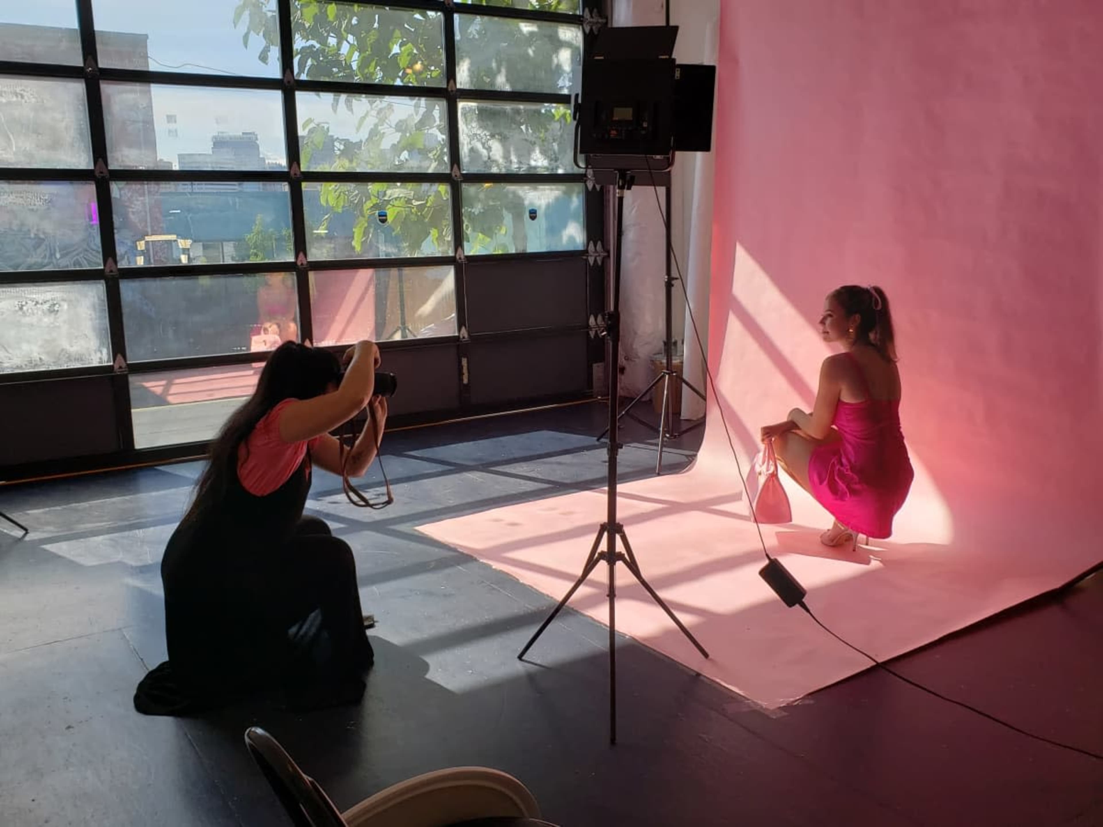A photographer captures images of a model posing on a pink backdrop in a well-lit studio.