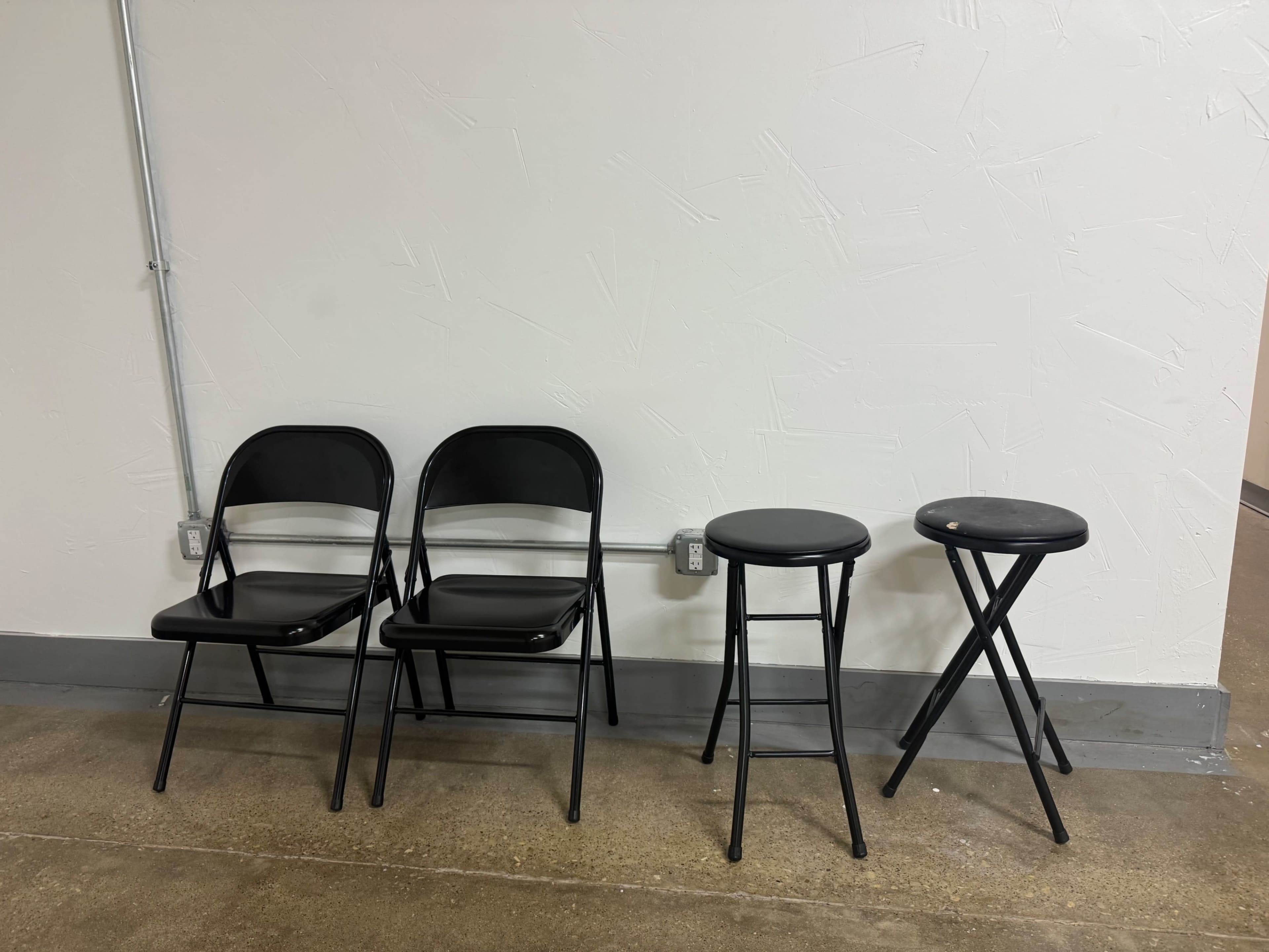 Four black folding chairs are positioned against a white wall, with three on the left and a black stool on the right.