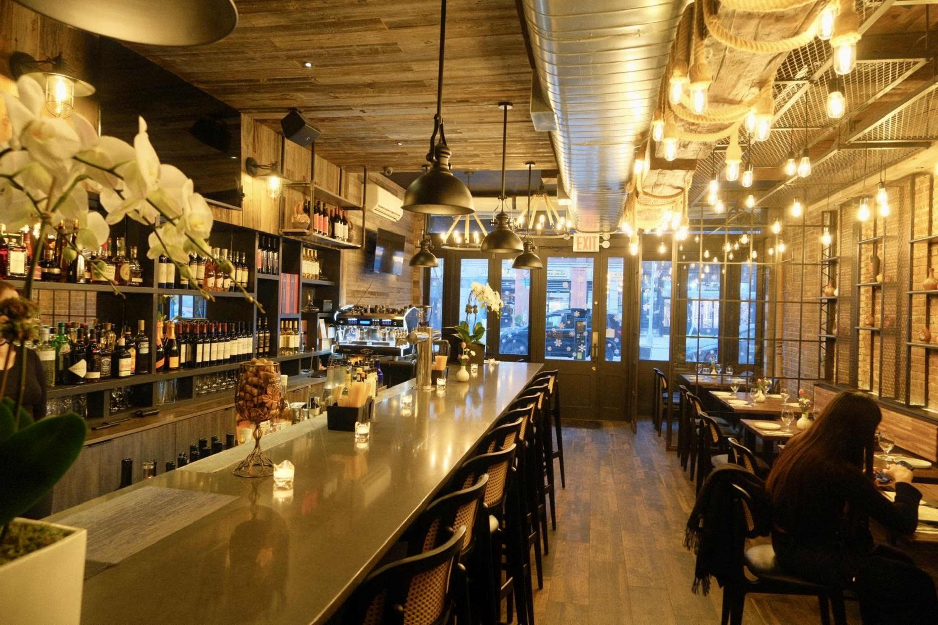 The image shows a warmly lit restaurant interior with a long bar, shelves filled with wine bottles, and a few diners seated at tables.