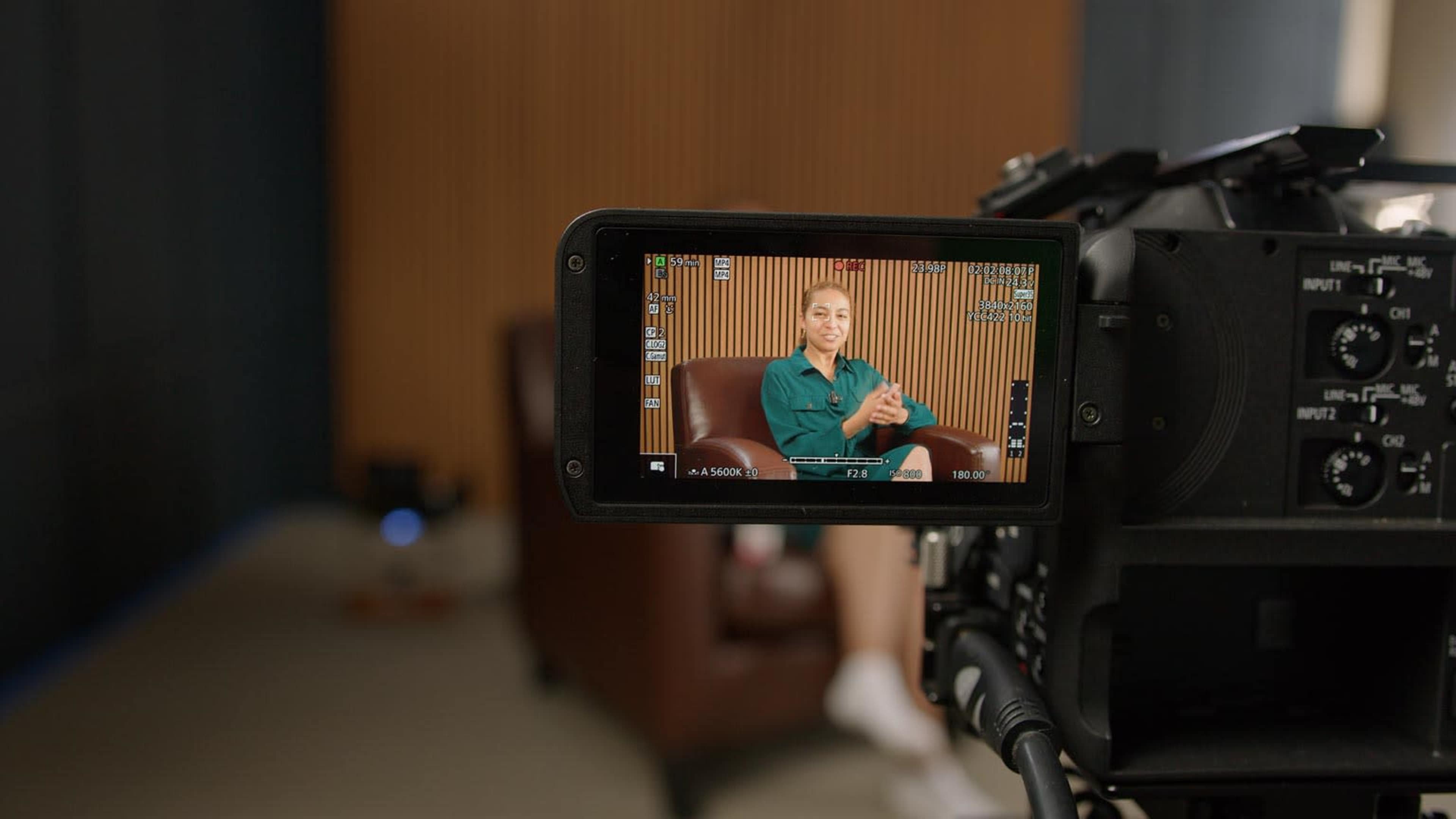 A camera is focused on a woman sitting in a chair, speaking against a wooden backdrop, while the camera's display shows her image.