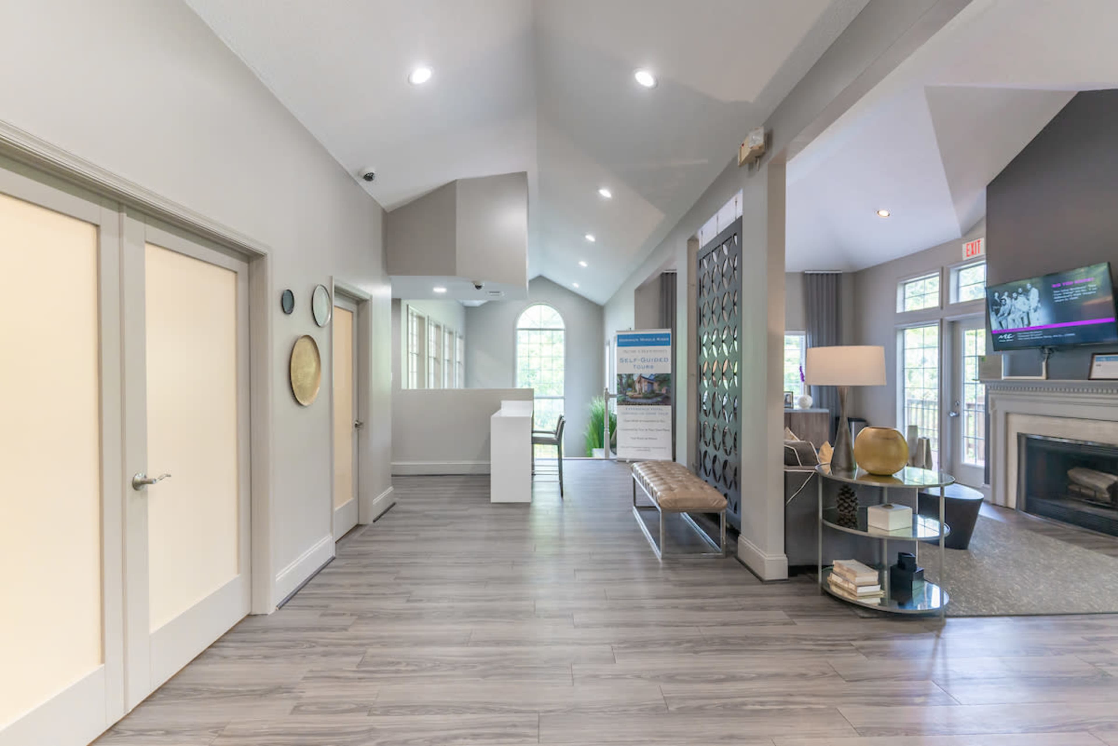 The image shows a modern lobby area featuring a high ceiling, gray walls, stylish furniture, and a large window providing natural light.
