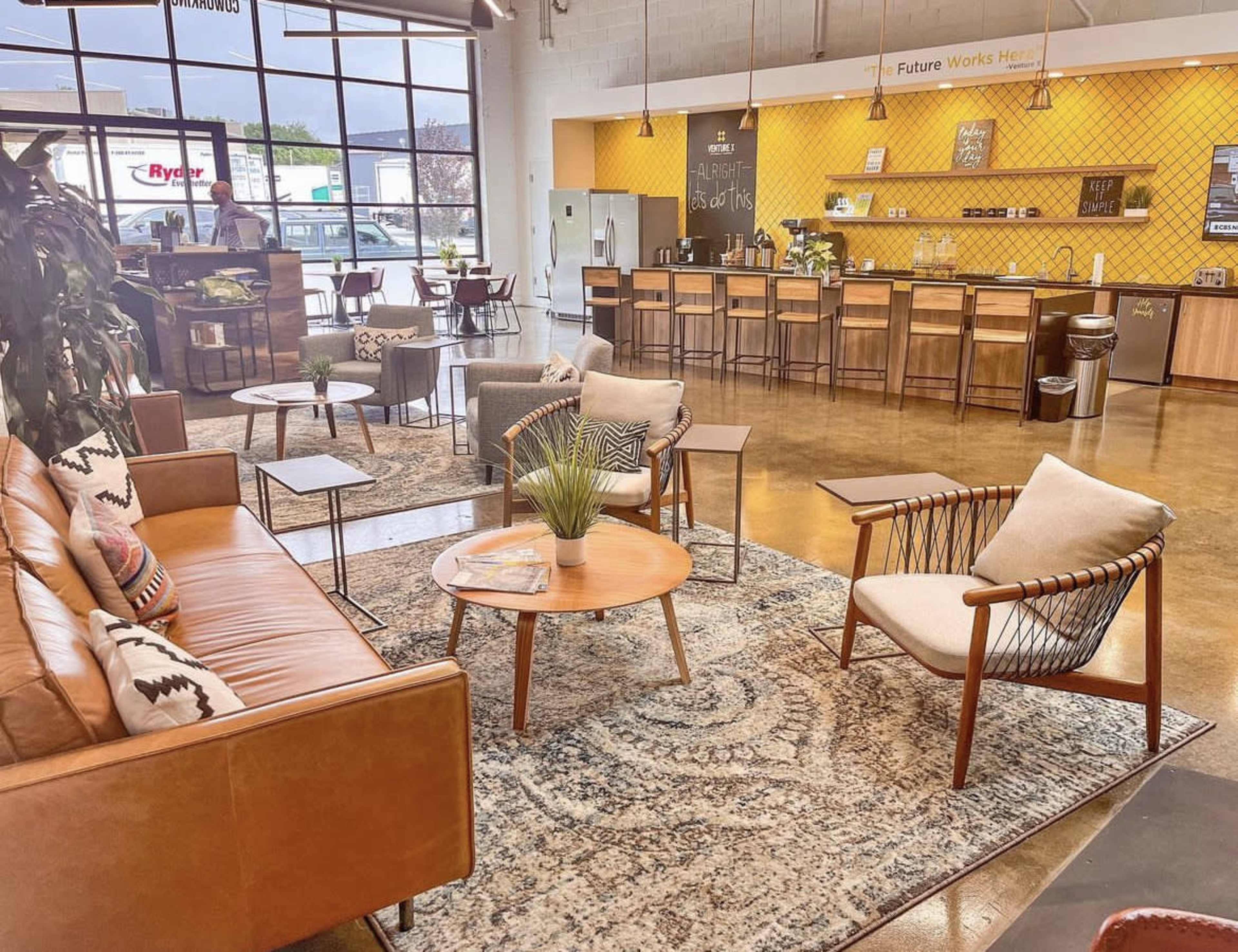 A cozy lounge area with brown leather sofas, wooden coffee tables, and a kitchenette in a bright, open space featuring large windows.