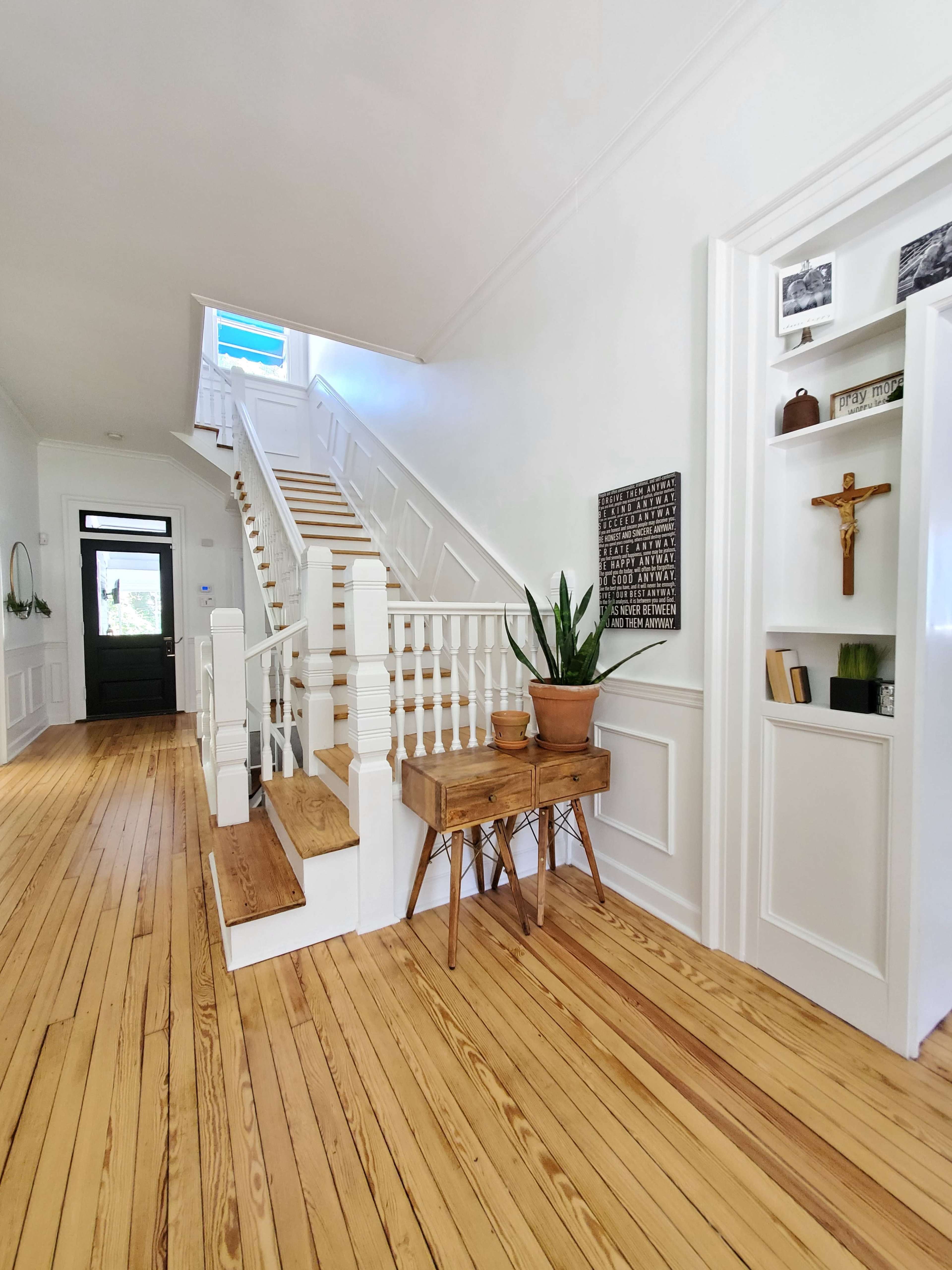 The image shows a well-lit hallway with wooden floors, featuring a staircase on one side and a small table with a potted plant on the other.