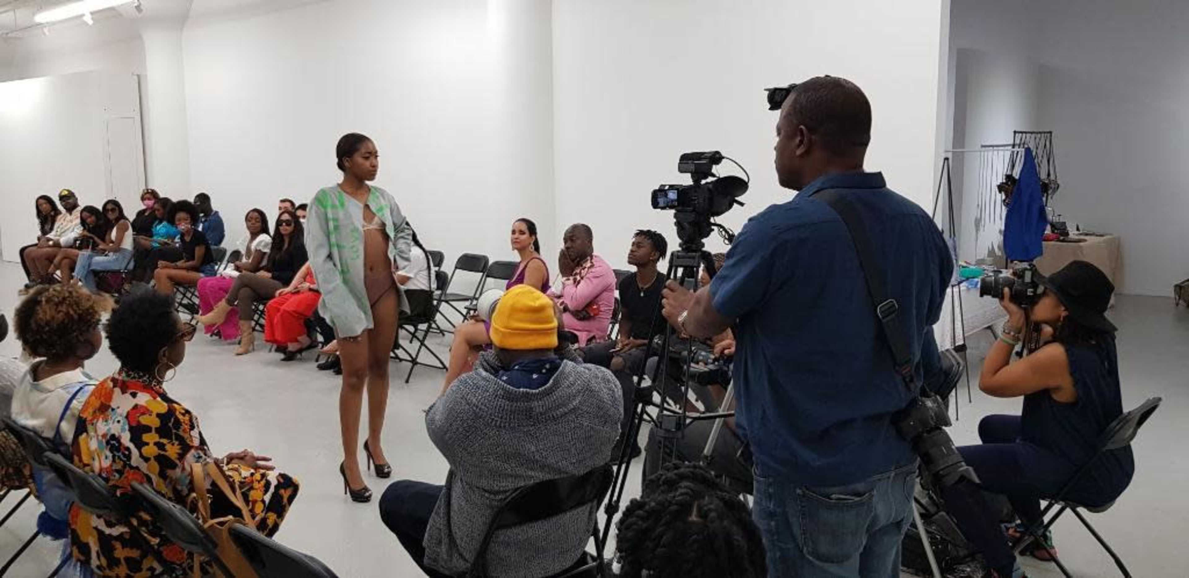 A model walks down a runway during a fashion event while photographers and attendees observe.