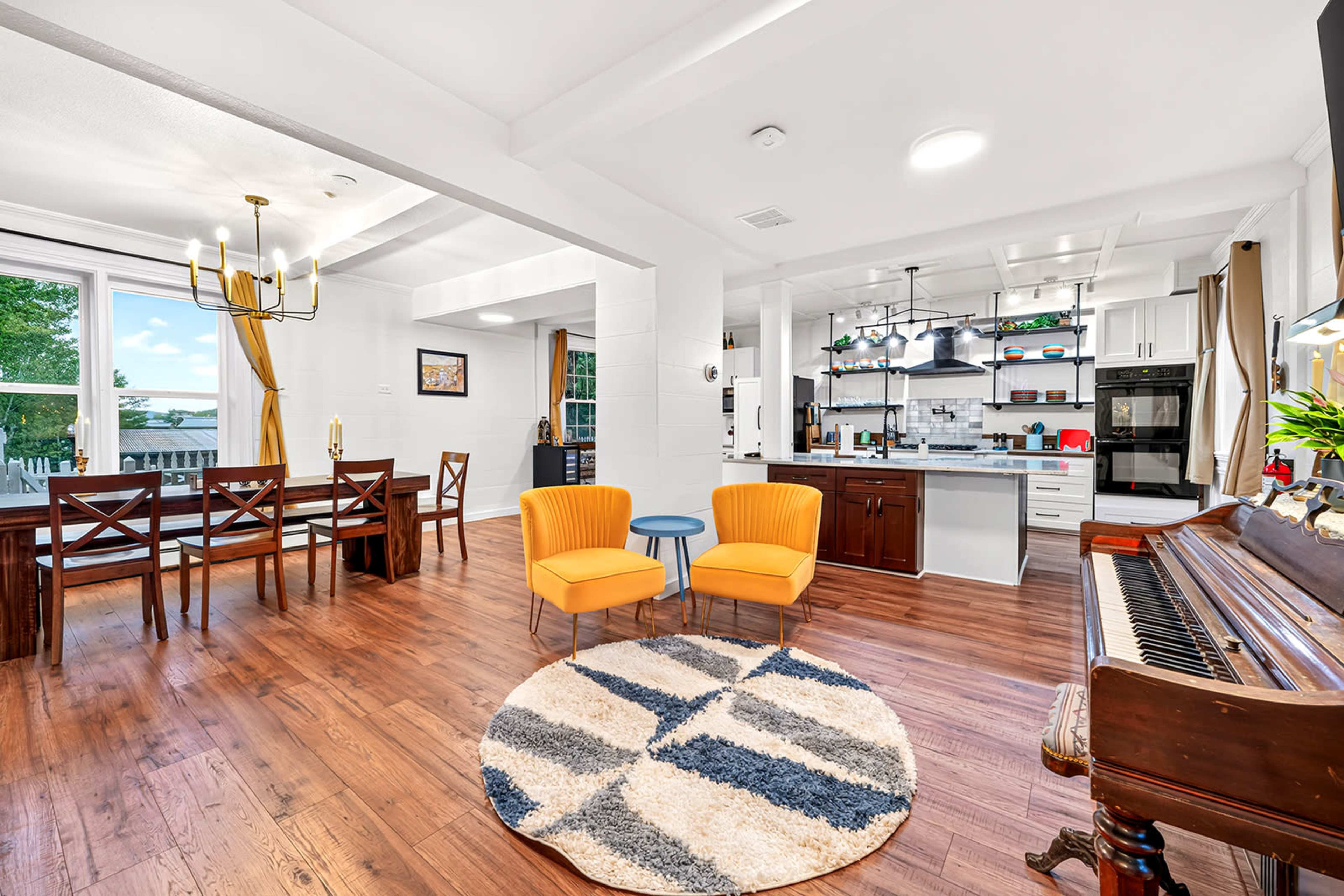 The image shows a bright, open-concept living and dining area featuring a piano, yellow chairs, a round patterned rug, a dining table, and a modern kitchen in the background.