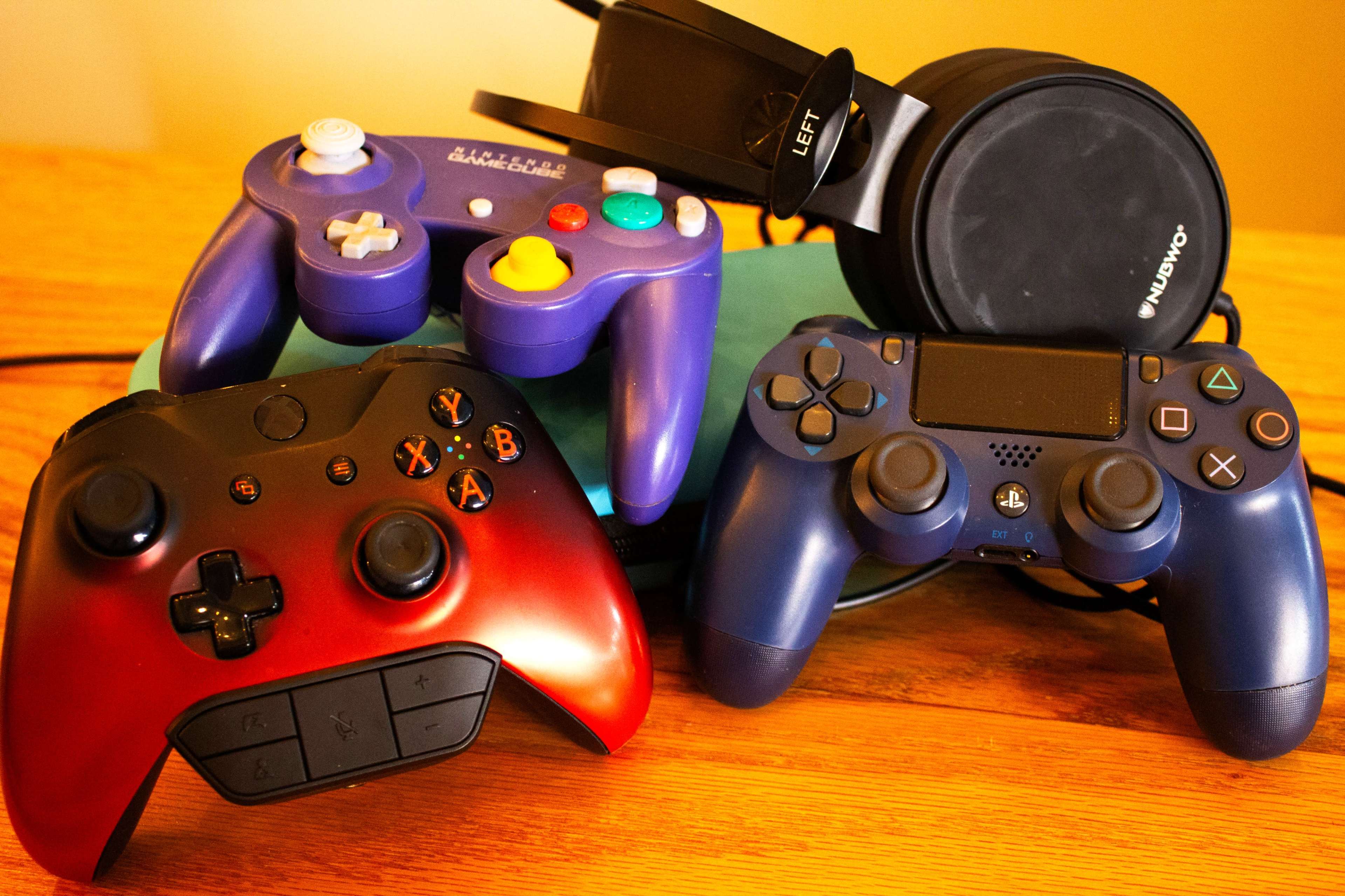The image shows a collection of gaming controllers in various colors stacked next to a pair of headphones on a wooden surface.