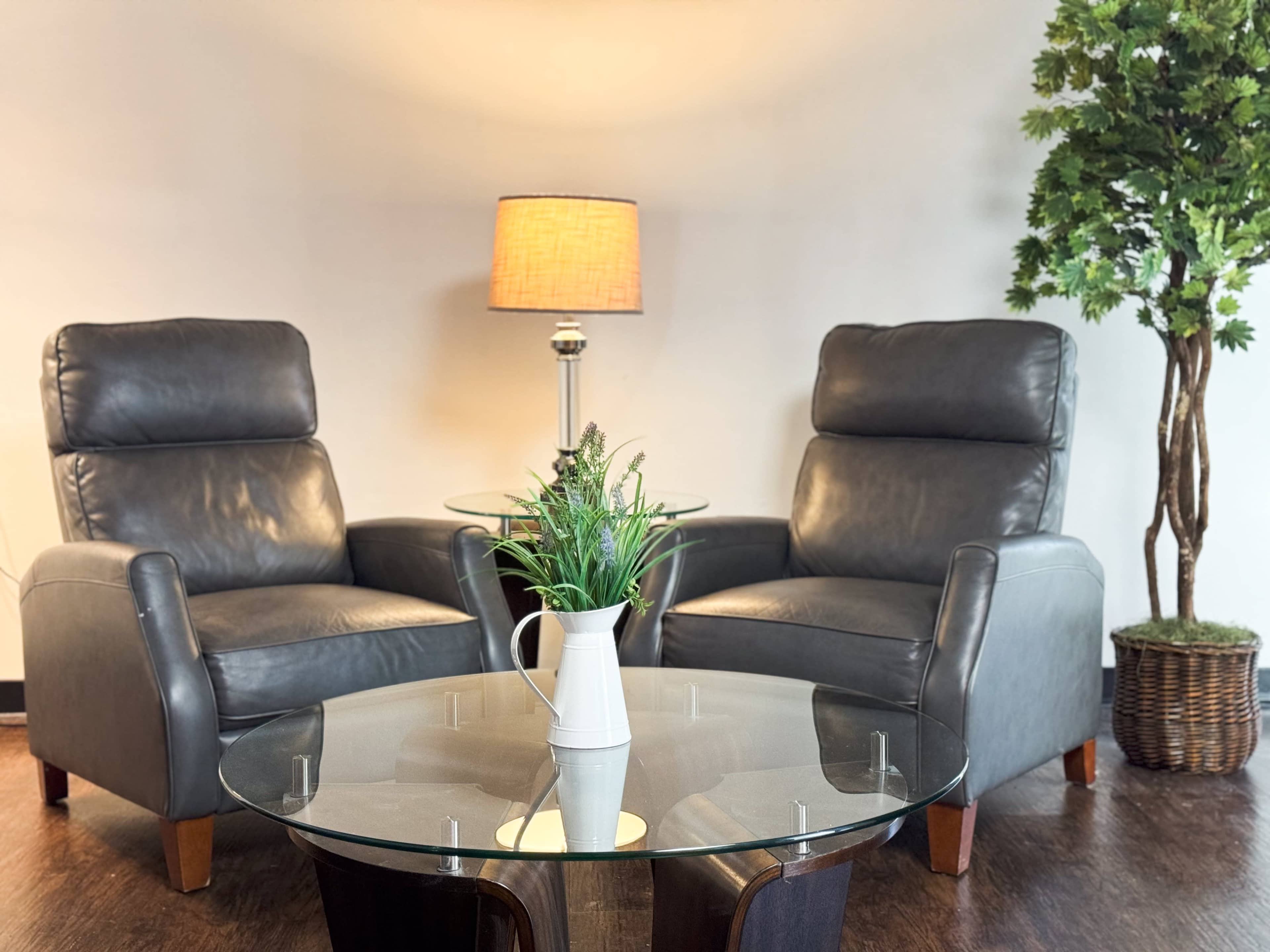 The image depicts two gray leather chairs facing each other across a glass coffee table, with a lamp and a potted plant nearby.