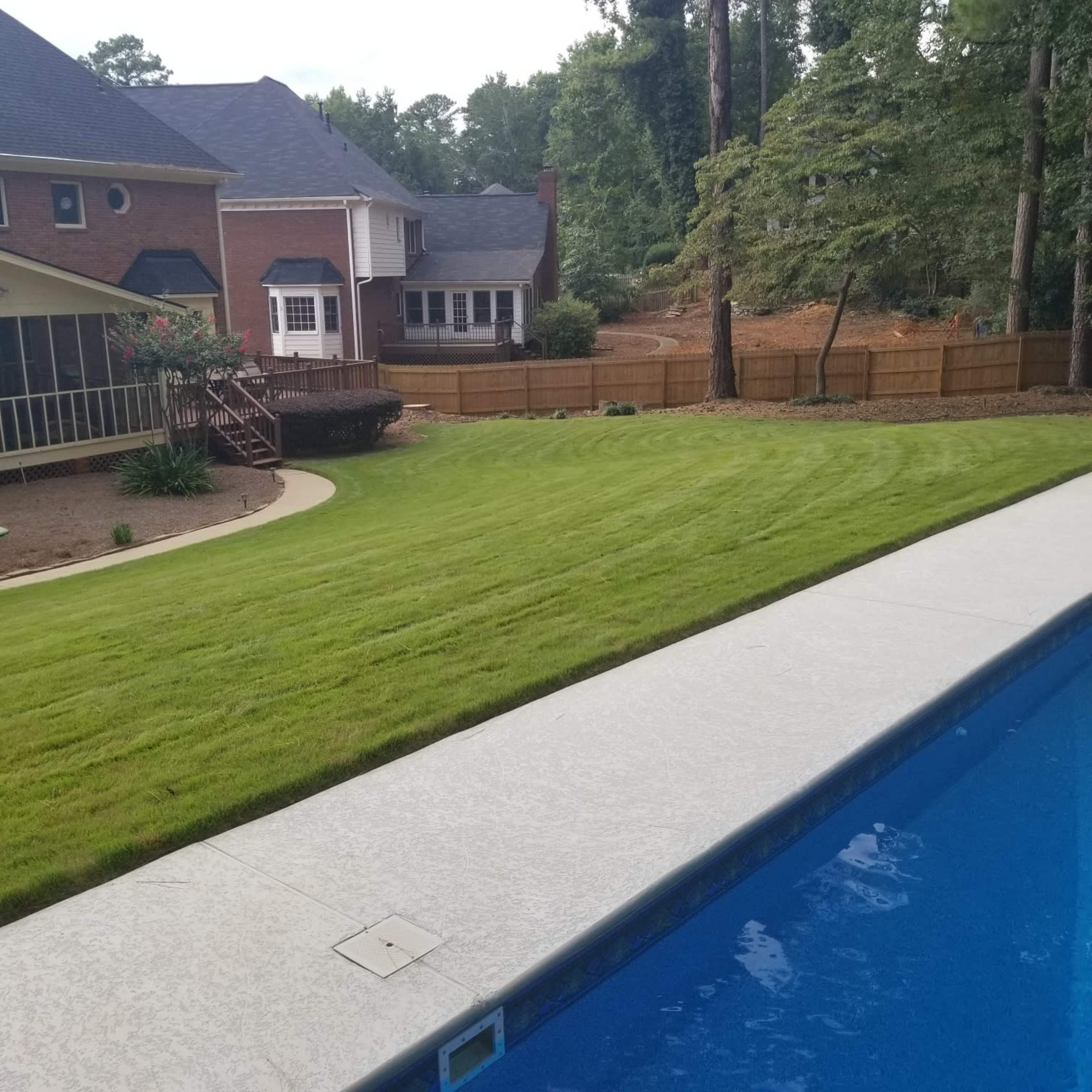 A neatly maintained lawn borders a swimming pool, with a fenced backyard and a two-story house in the background.