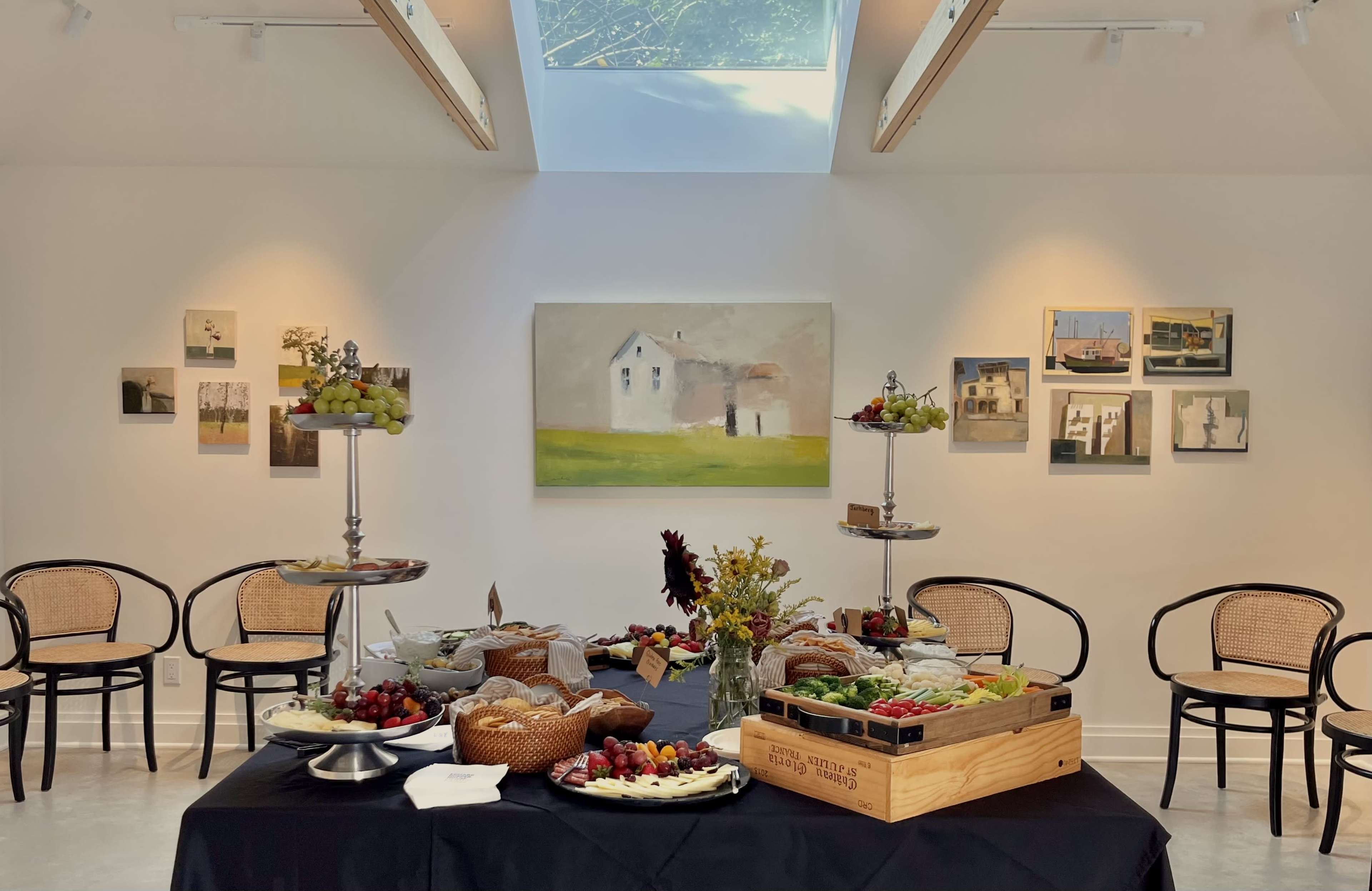 A well-decorated dining space features a black tablecloth with an assortment of fruits and pastries, surrounded by elegant chairs and a large artwork on the wall.