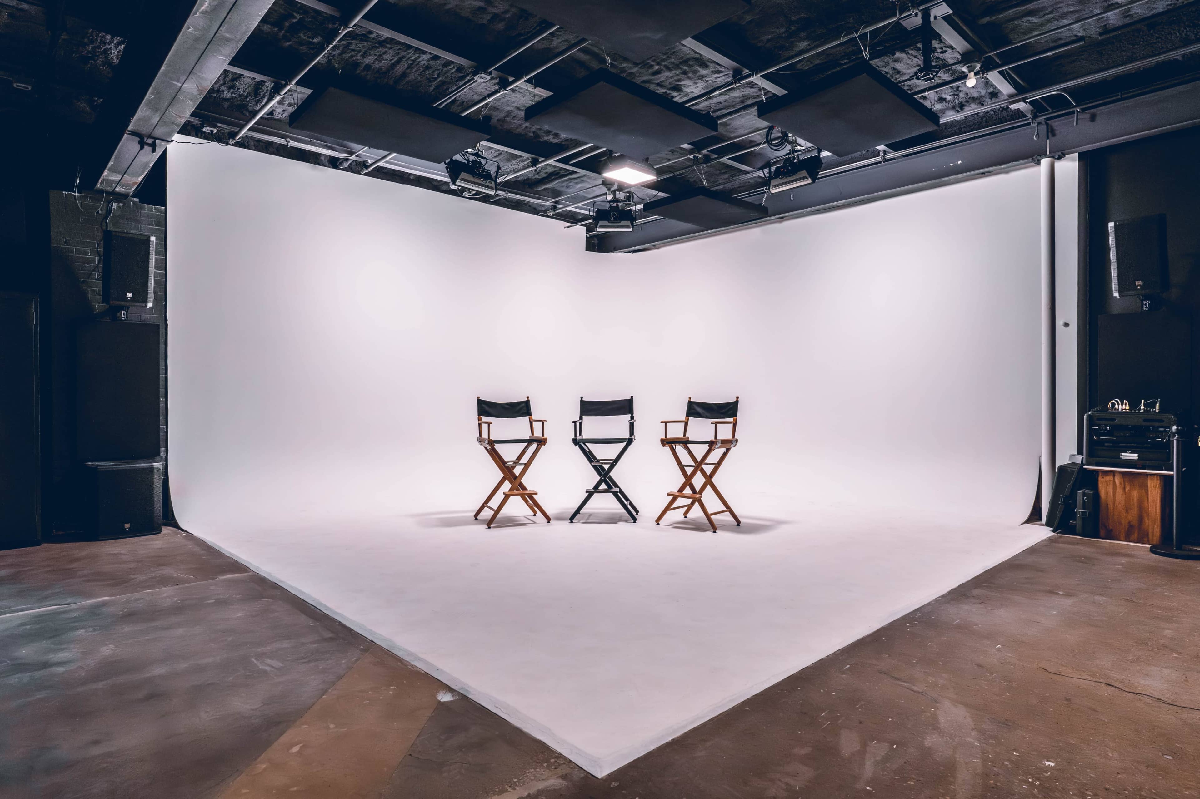 The image shows an empty studio space with three wooden director's chairs arranged in a row against a blank white backdrop.