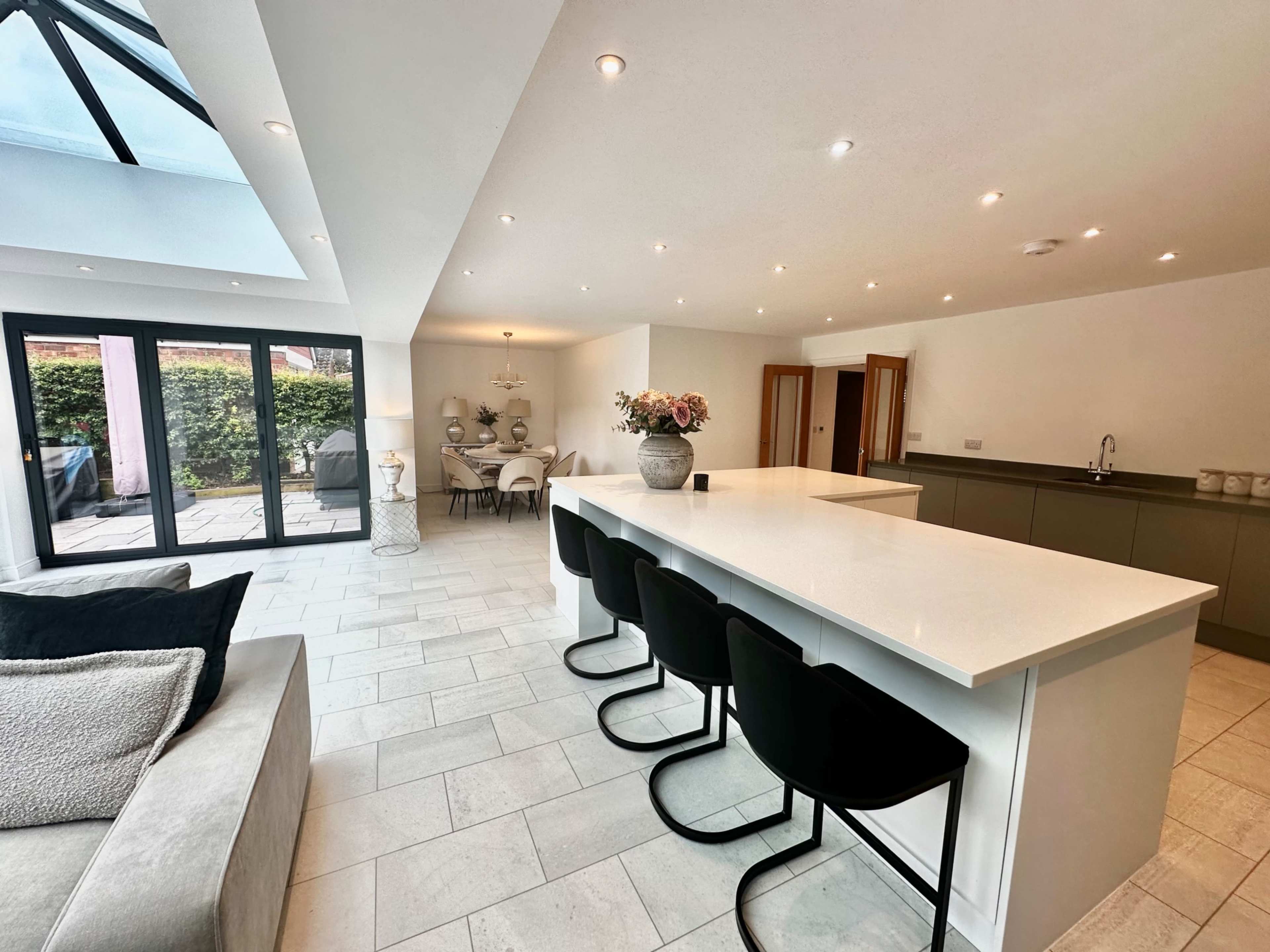 A modern open-plan kitchen and living area with a large island, black bar stools, and a dining space visible in the background.