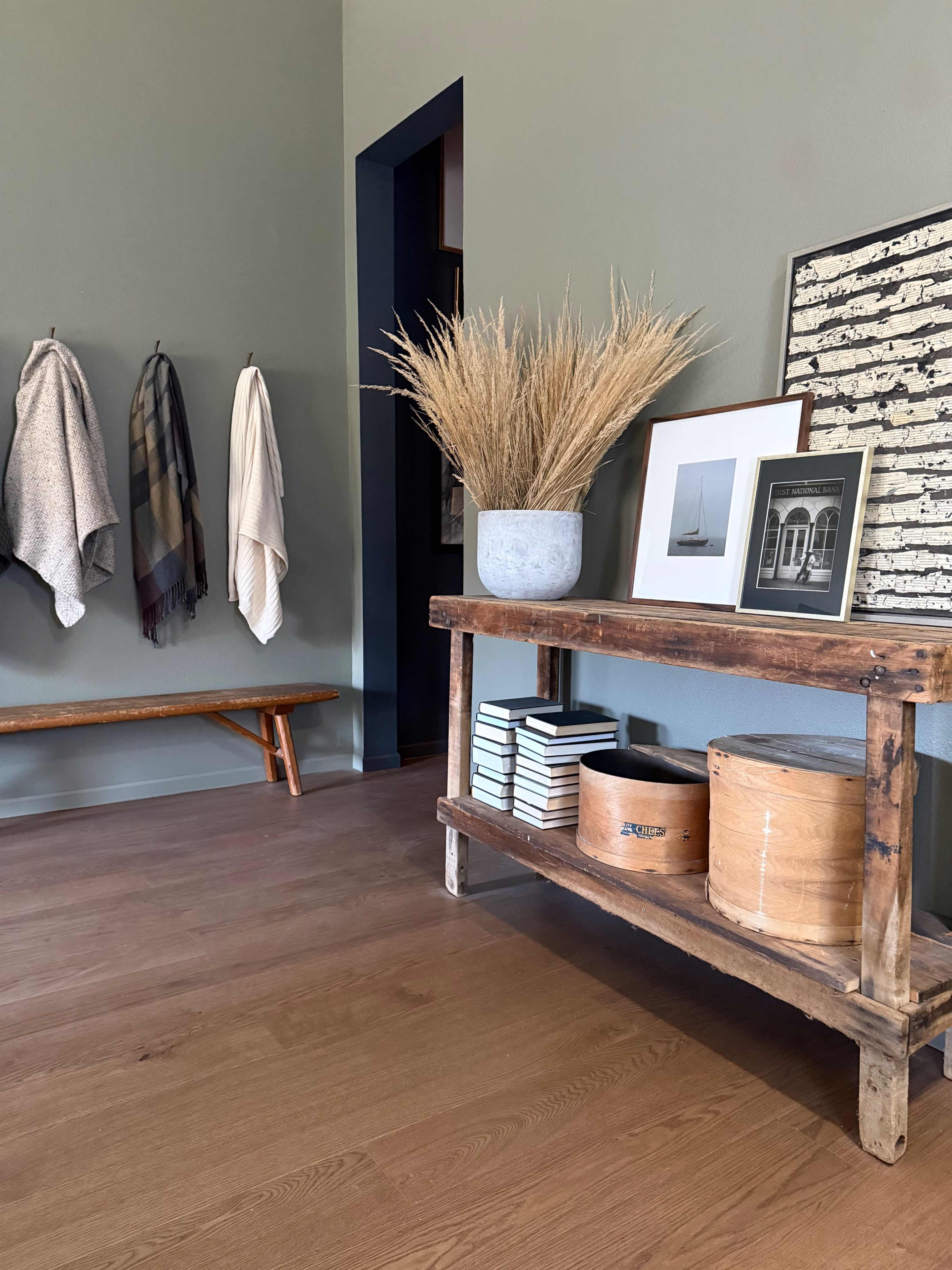 The image features a modern entryway with wooden flooring, a storage bench, a table displaying decorative items, and wall-mounted hooks with hanging blankets.