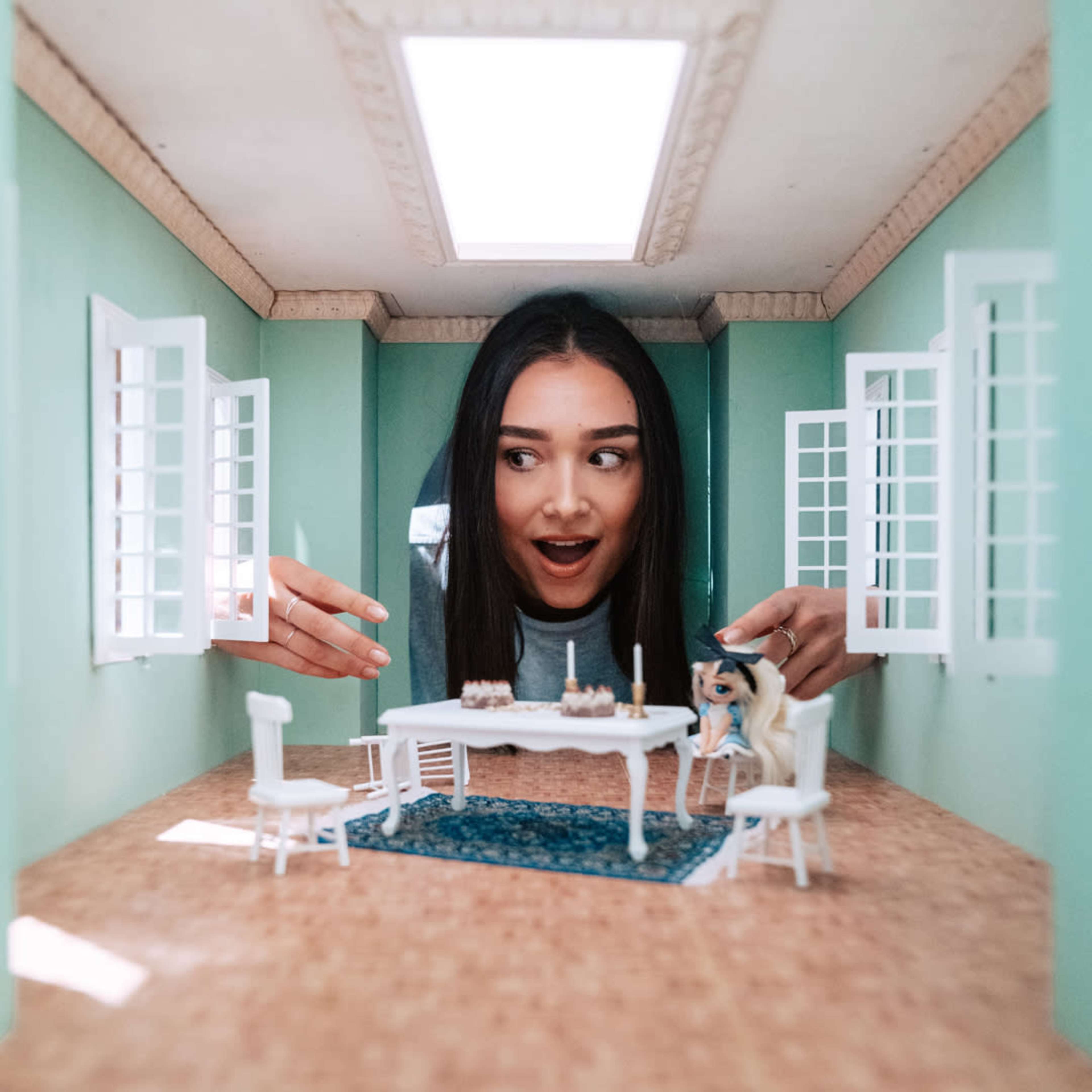 A young woman interacts with a dollhouse, placing a doll at a miniature dining table set within the colorful room.