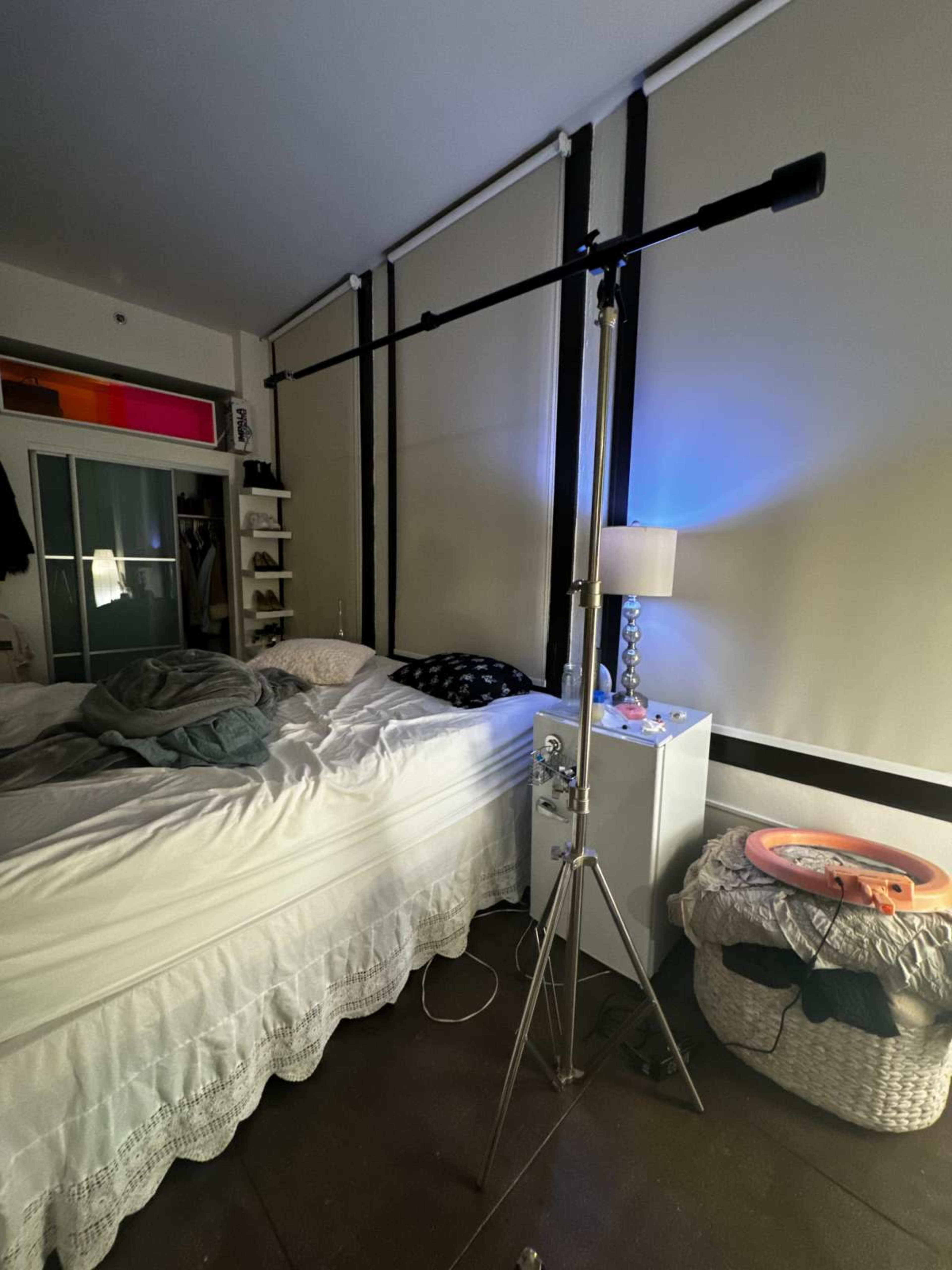 The image shows a bedroom with a bed covered in a gray blanket, a lamp on a nightstand, and a lighting stand positioned near the bed.