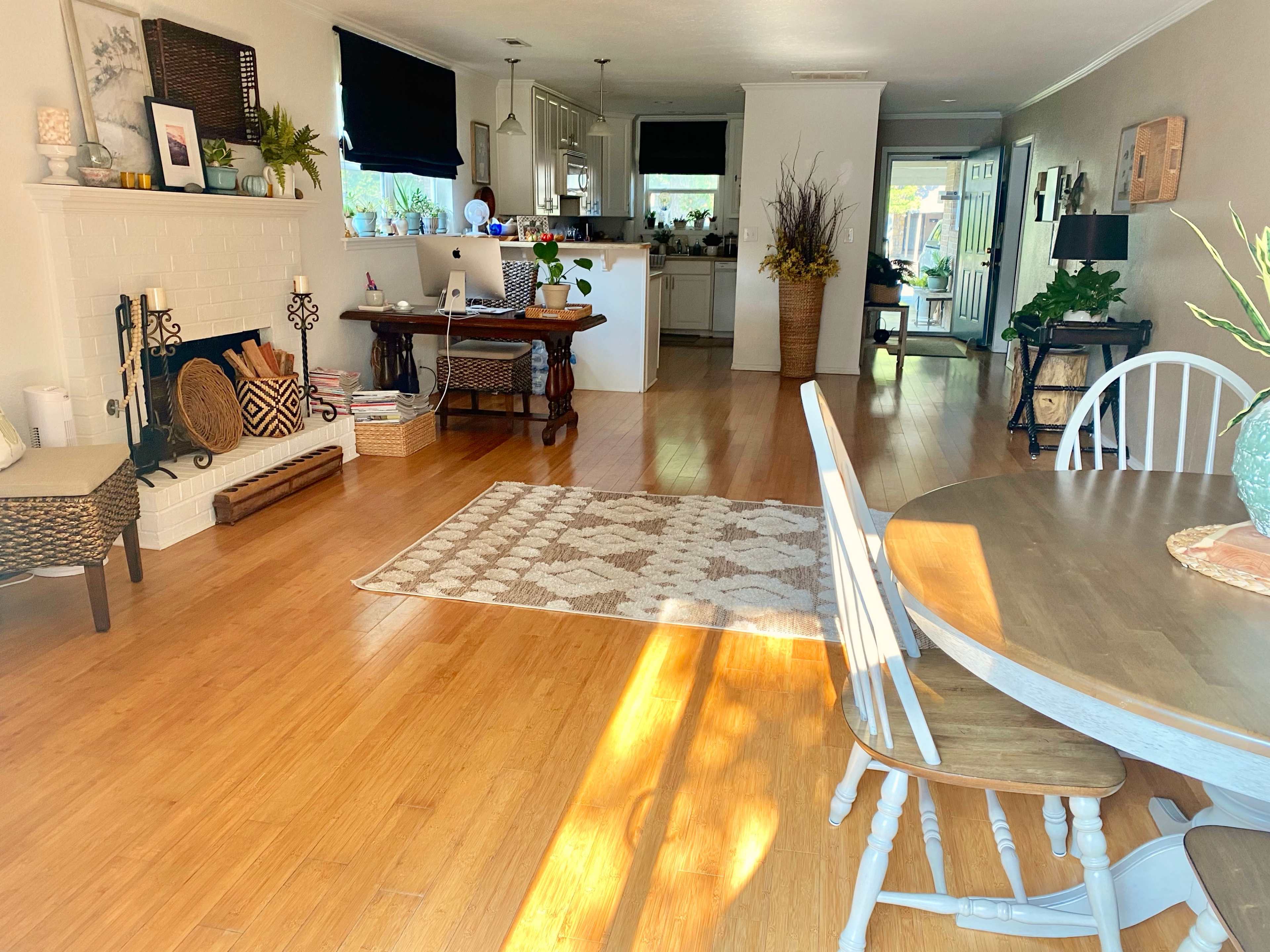 The image shows a bright, open living area featuring a dining table, a rug, a small fireplace, and a view of a kitchen in the background.