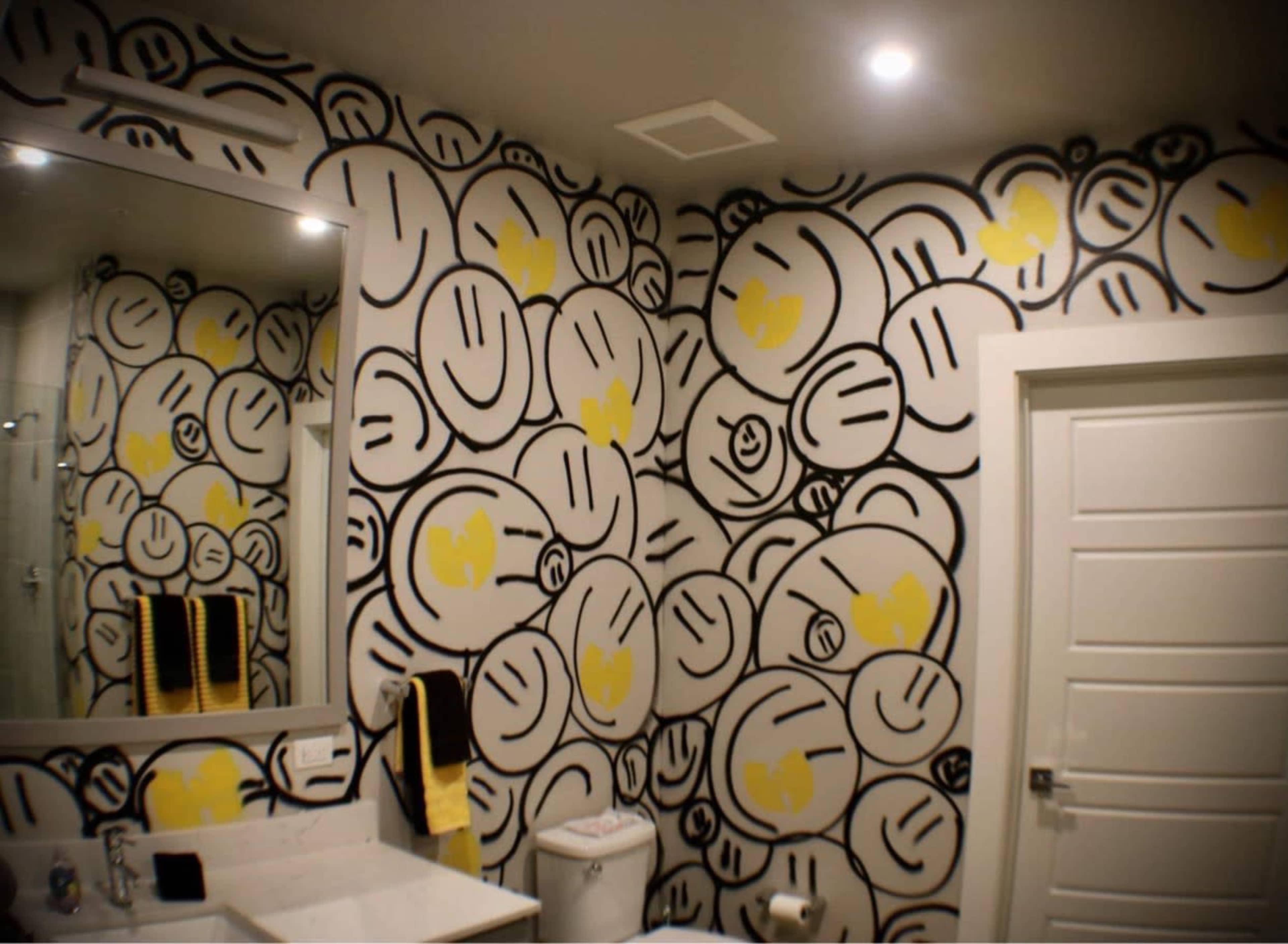 The bathroom features walls covered in a playful black and yellow smiley face mural, with a white sink, modern mirror, and towel rack.