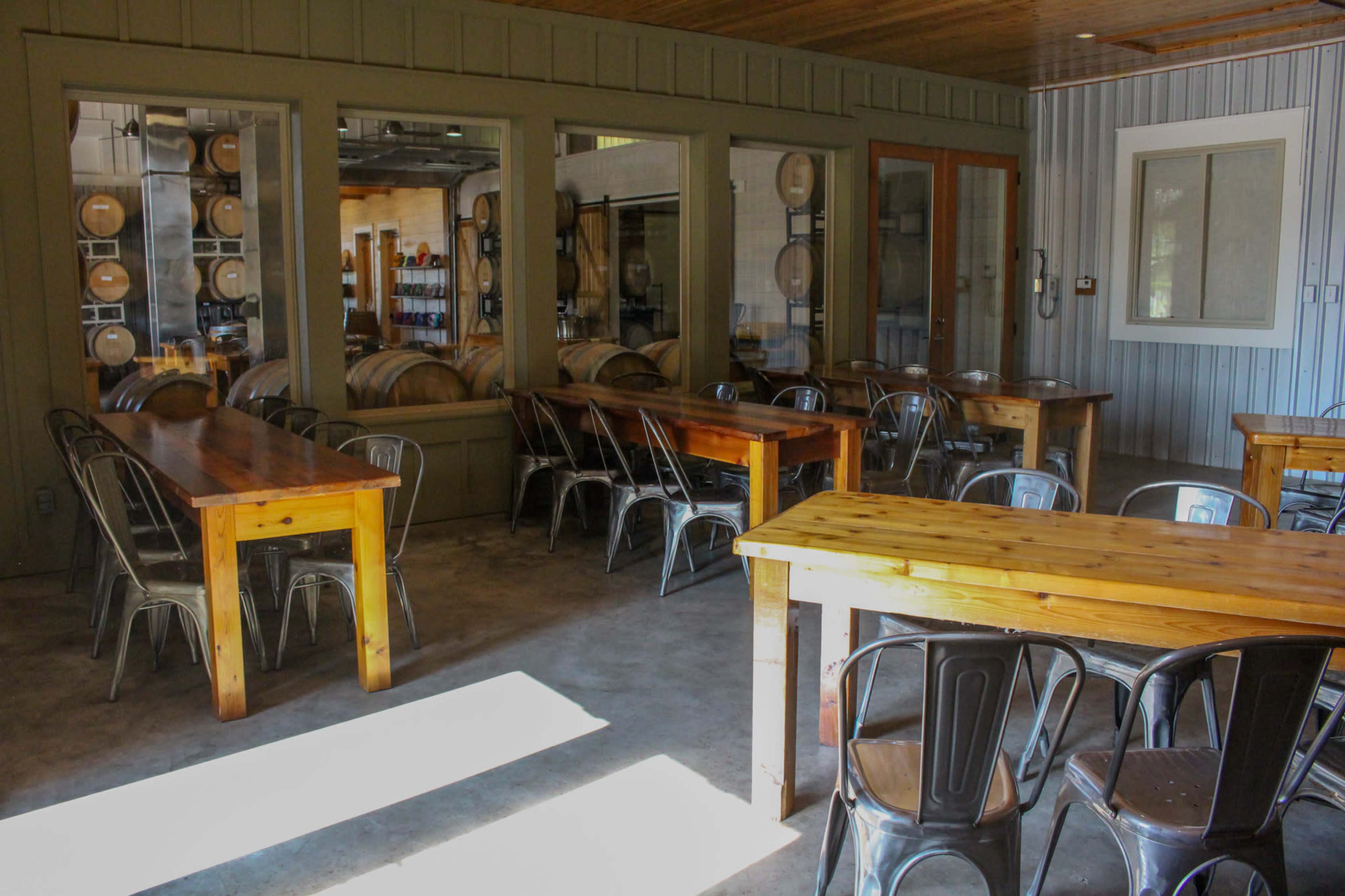 A dining area with wooden tables and metal chairs, alongside a view of storage barrels through large windows.