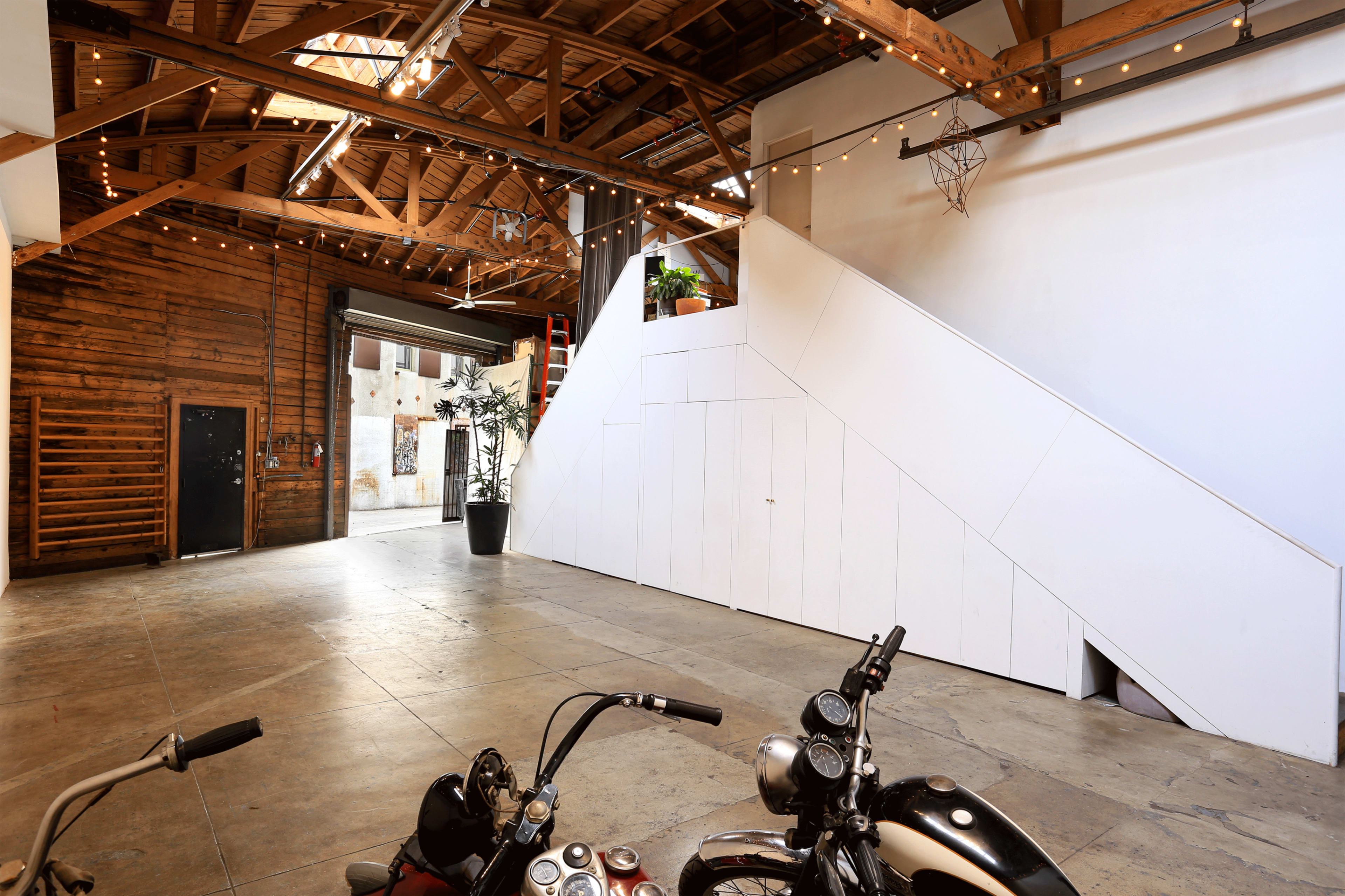 The image shows an industrial-style interior space with wooden beams, concrete flooring, and two motorcycles parked in the foreground.