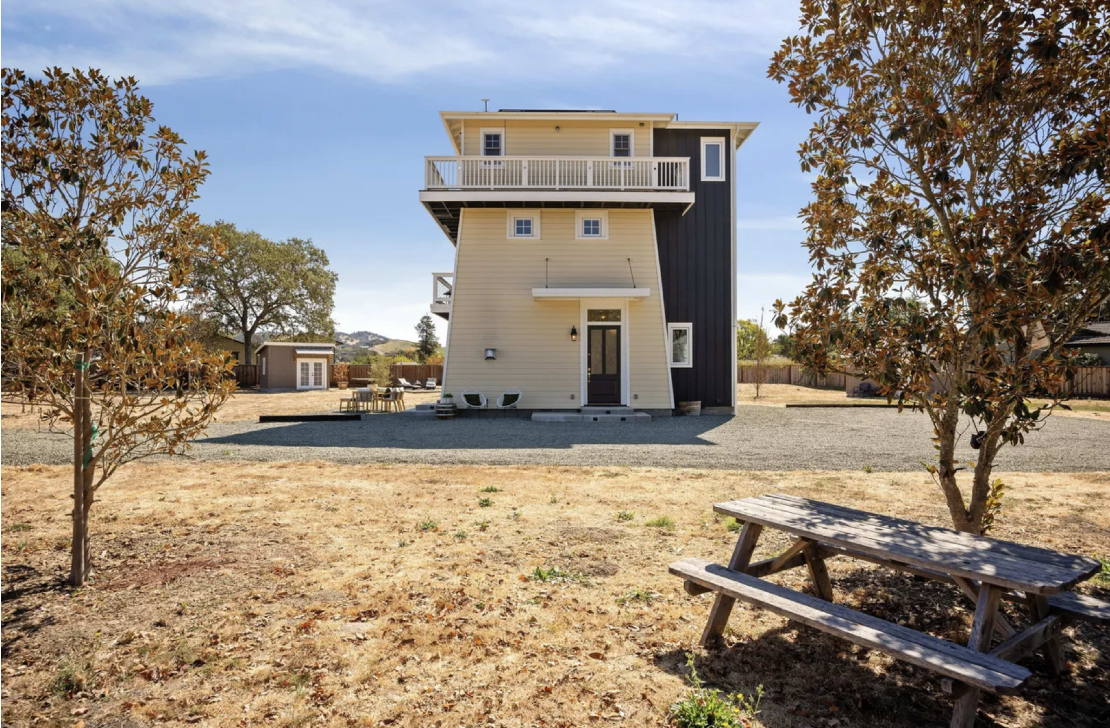 The image shows a three-story house with a mix of yellow and dark siding, set in a dry yard with a picnic table and surrounded by trees.