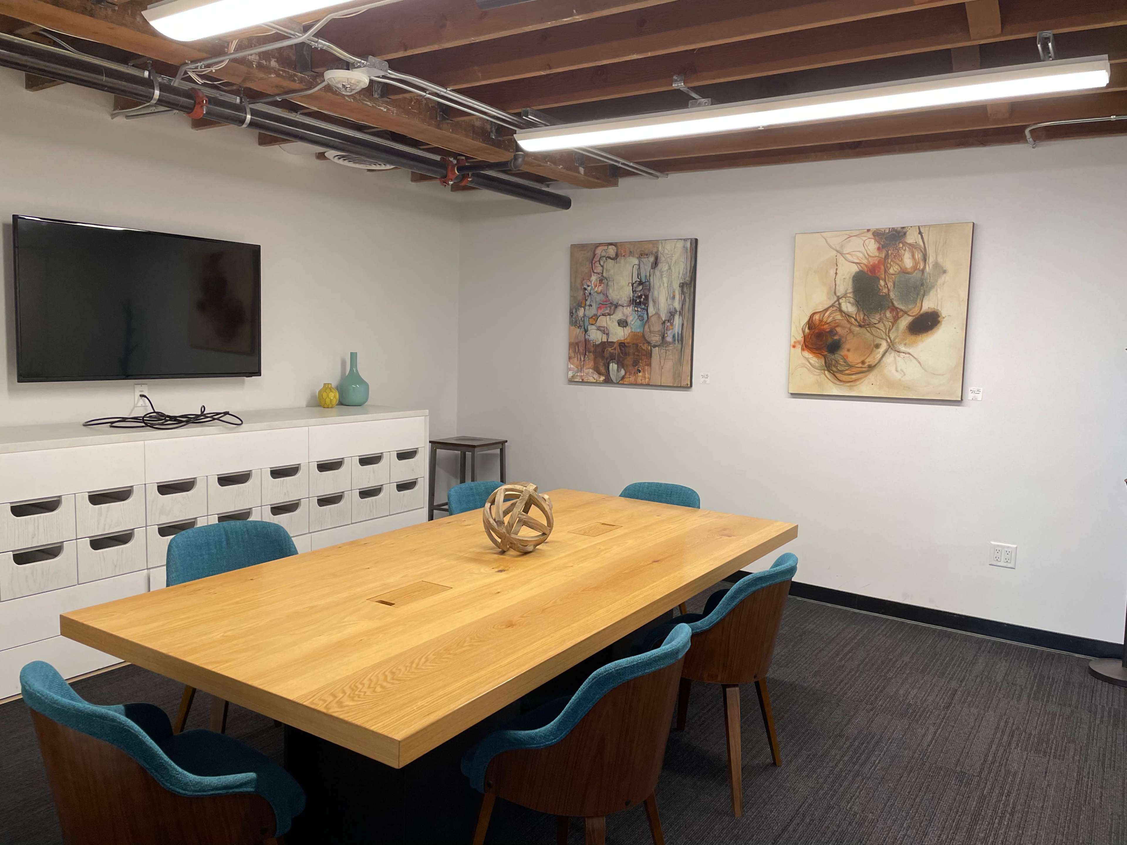 A conference room features a long wooden table surrounded by blue chairs, with a television mounted on the wall and two abstract art pieces hanging nearby.