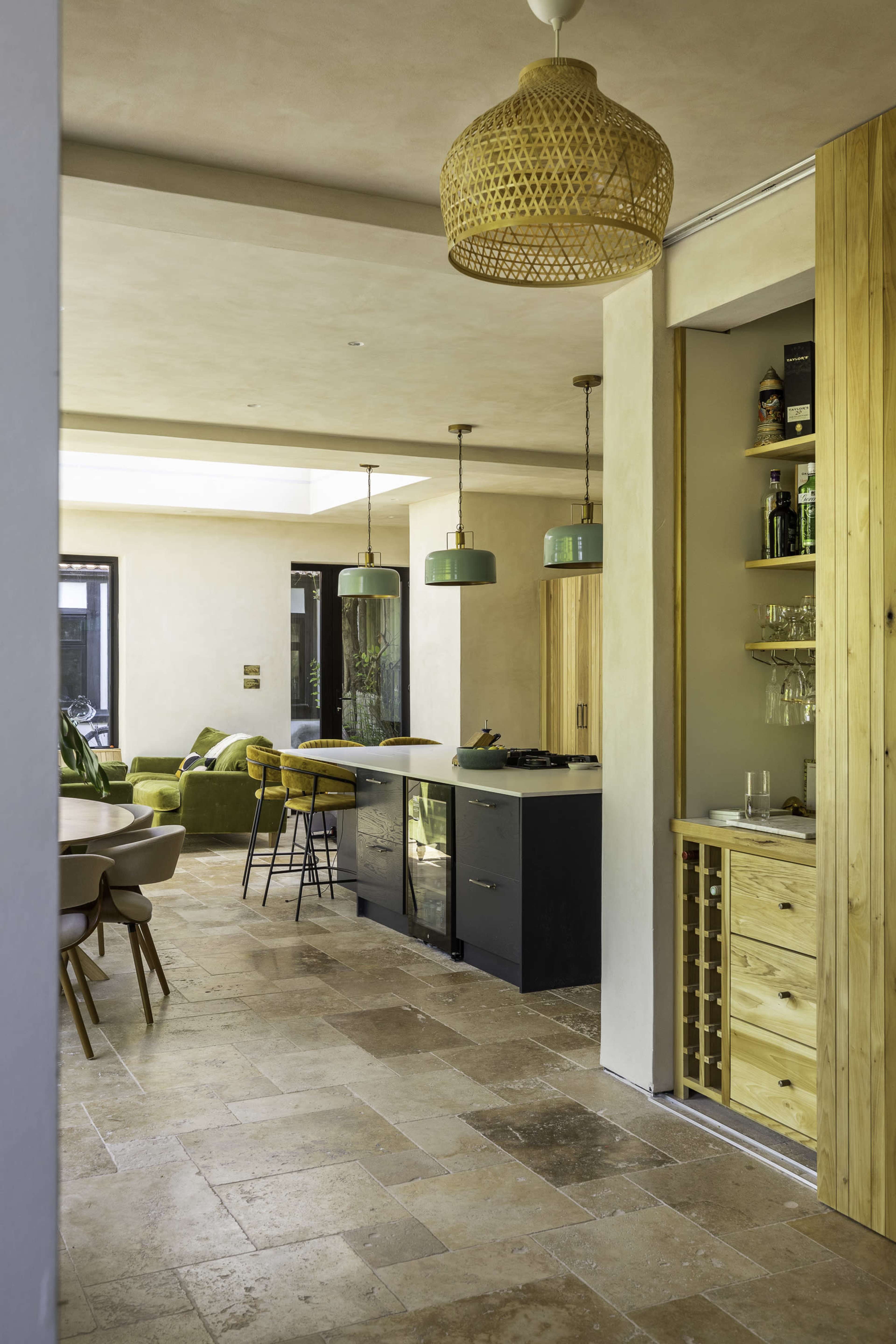 A modern kitchen with earthy tones features a central island, wooden cabinetry, pendant lights, and a dining area with a table and chairs.