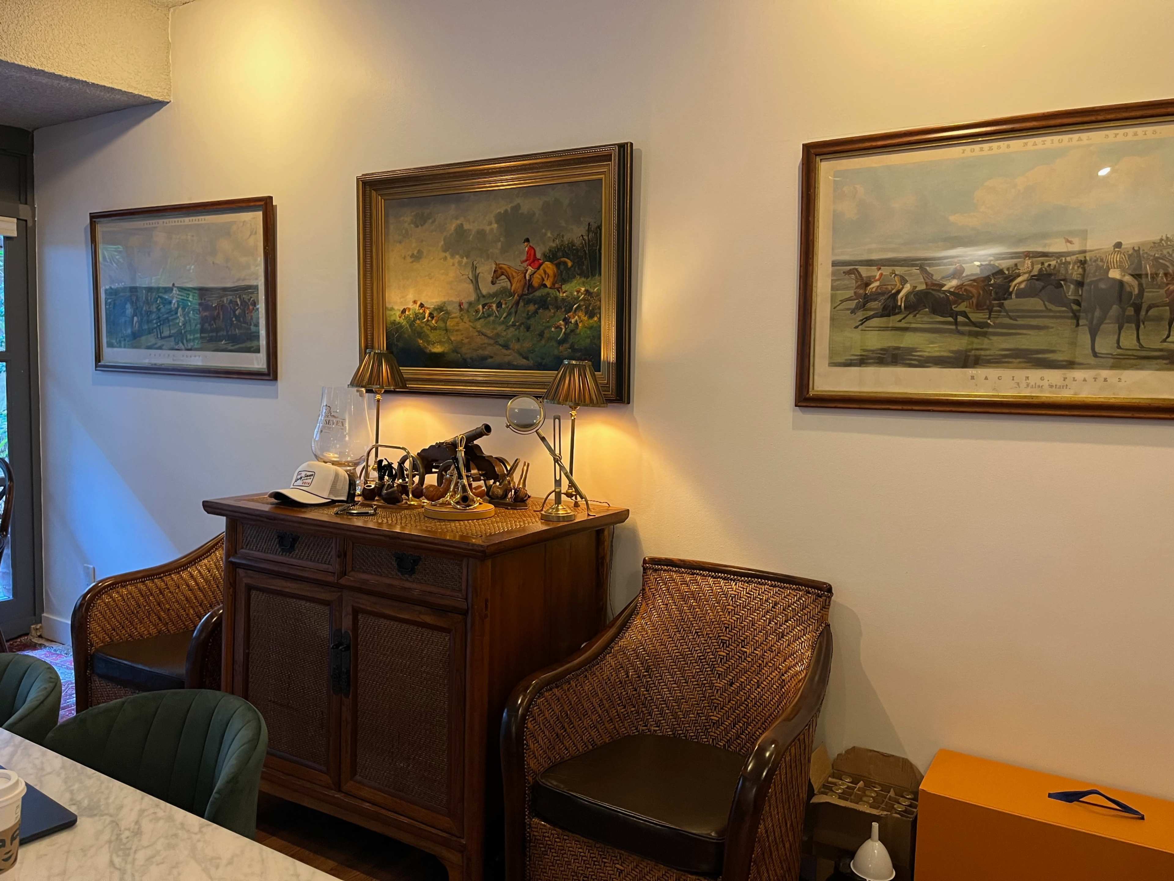 The scene shows an interior with two framed paintings of equestrian scenes hanging above a wooden sideboard, which is accompanied by two wicker chairs and a lamp.