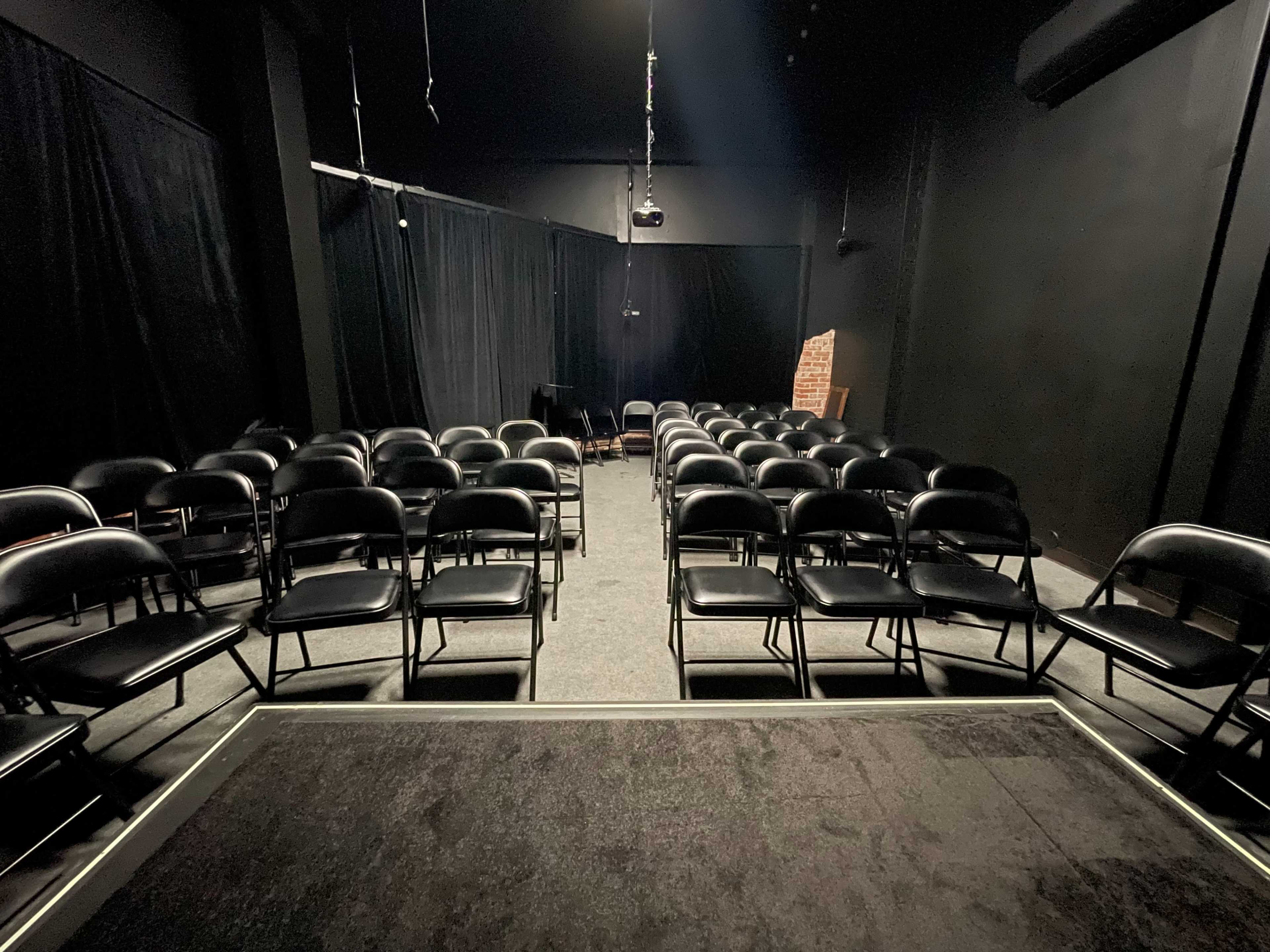 A dimly lit indoor performance space with rows of black folding chairs facing a small stage, surrounded by black curtains.