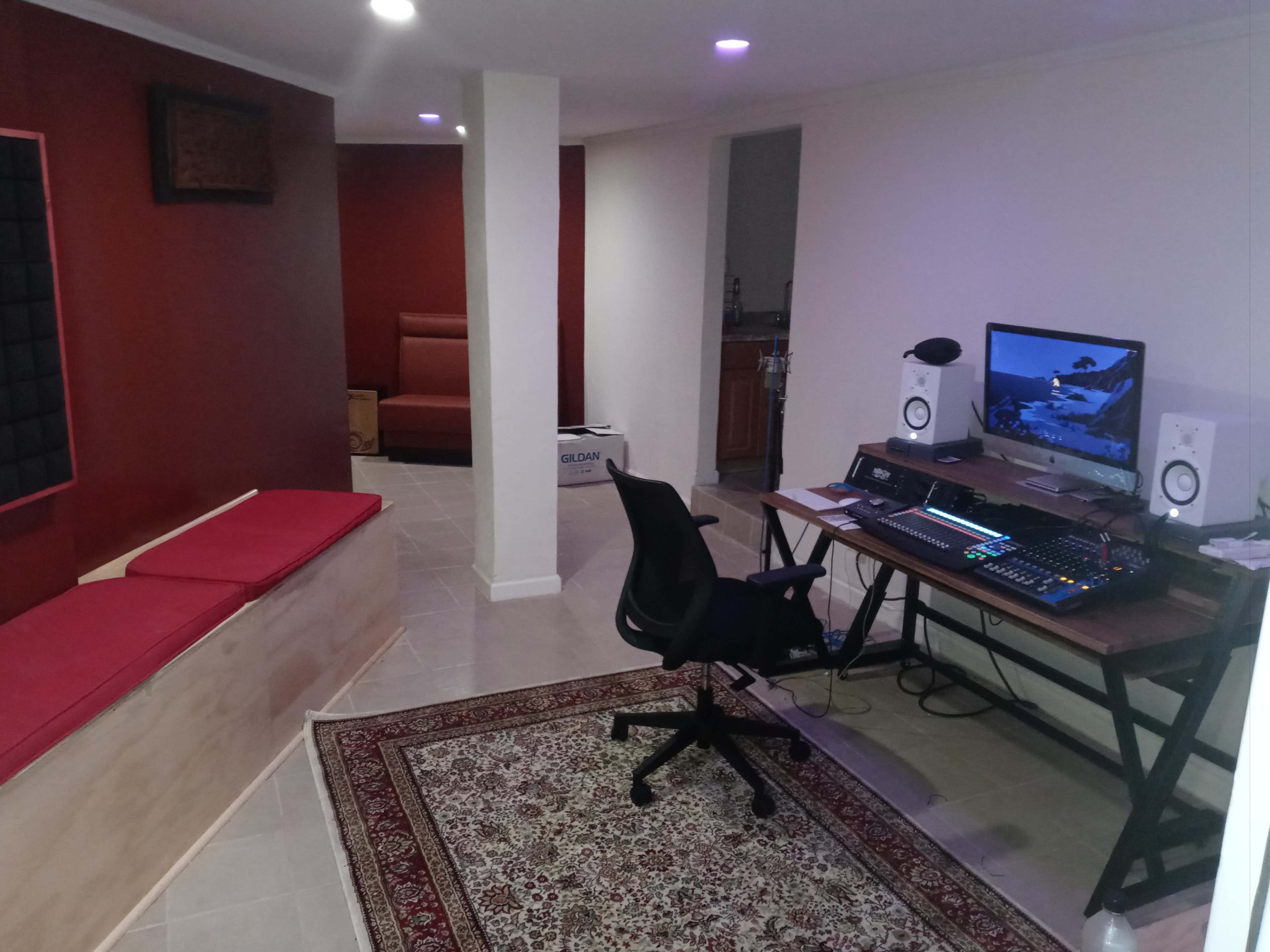 The image shows a basement space with a seating area on the left, a computer desk with monitors and sound equipment on the right, and a patterned rug on the floor.