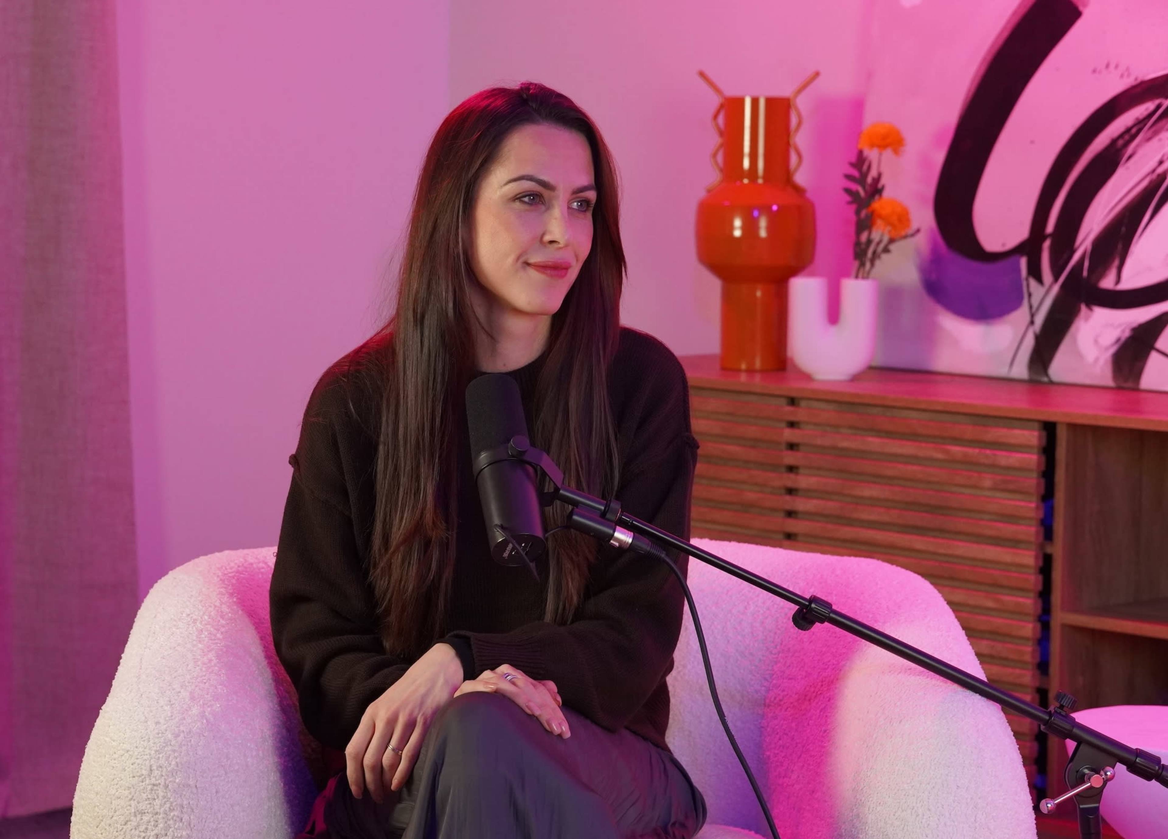 A woman is sitting in a white chair, speaking into a microphone, with colorful decor in the background.