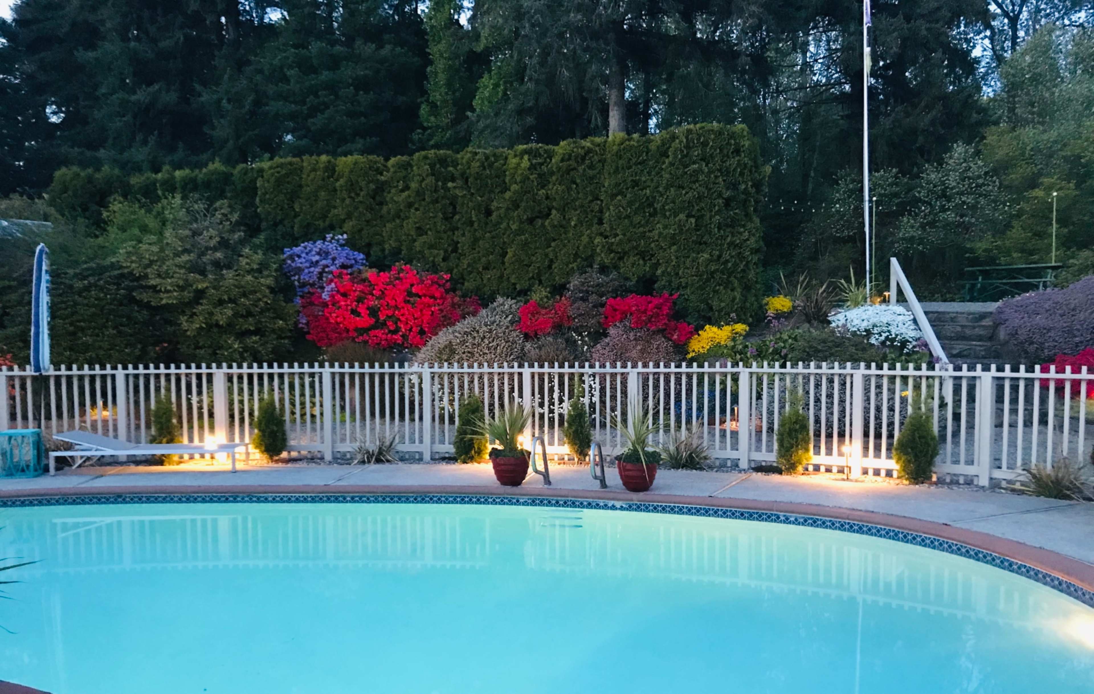A swimming pool surrounded by a white fence, with well-maintained hedges and colorful flower beds in the background.