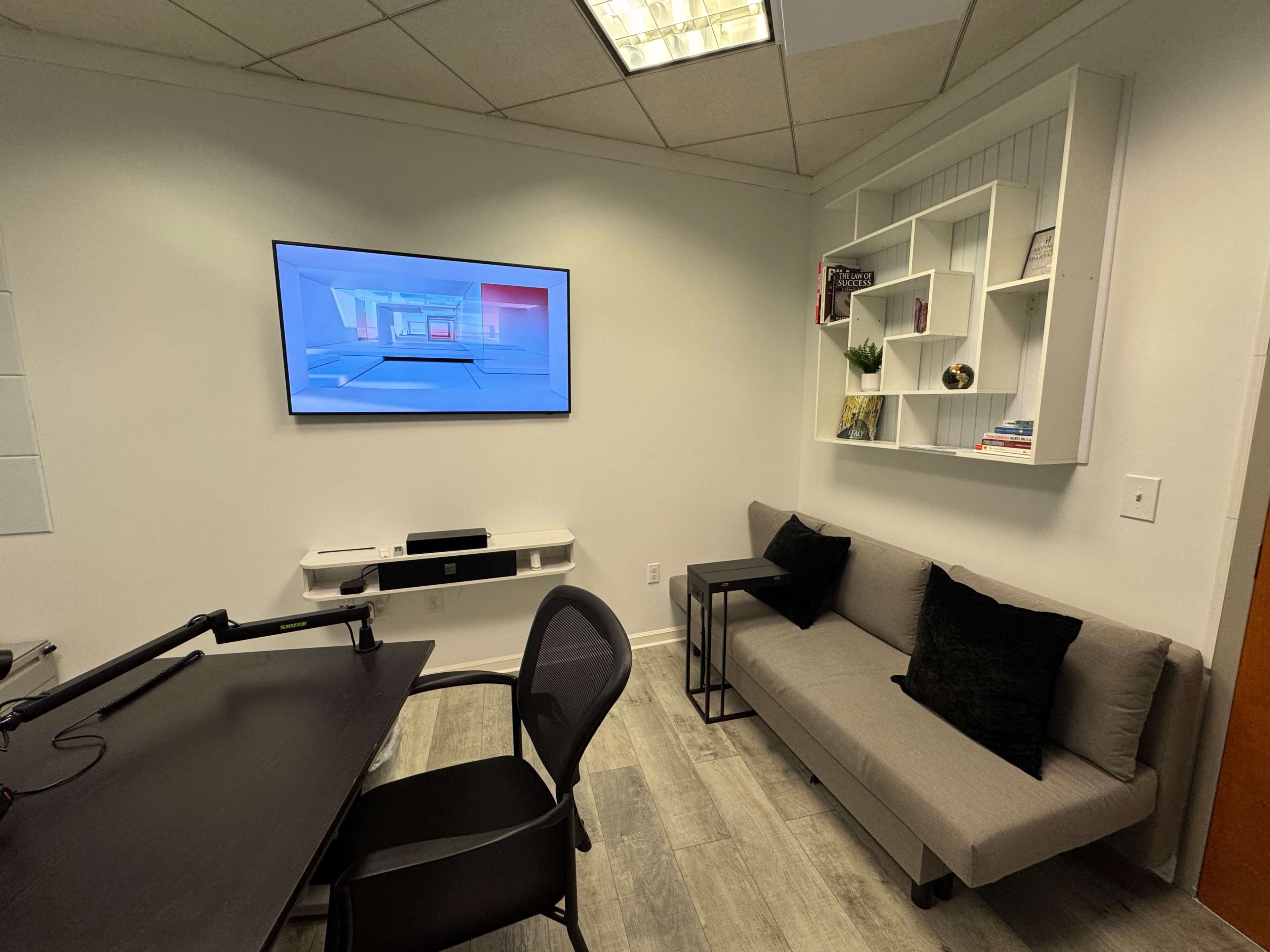 The image shows a small, modern office space with a wall-mounted television, a sofa, a desk with a chair, and shelves displaying various items.